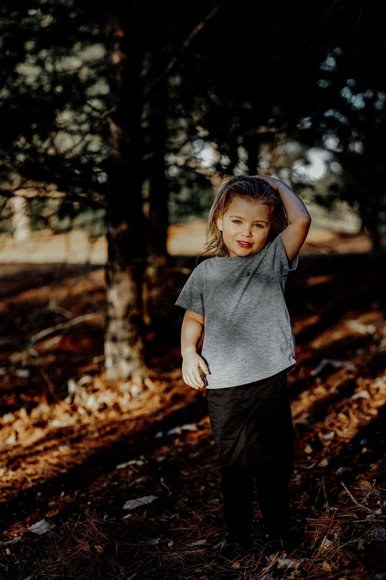 Playful young child in gray t-shirt posing with hand on head in dappled forest light.