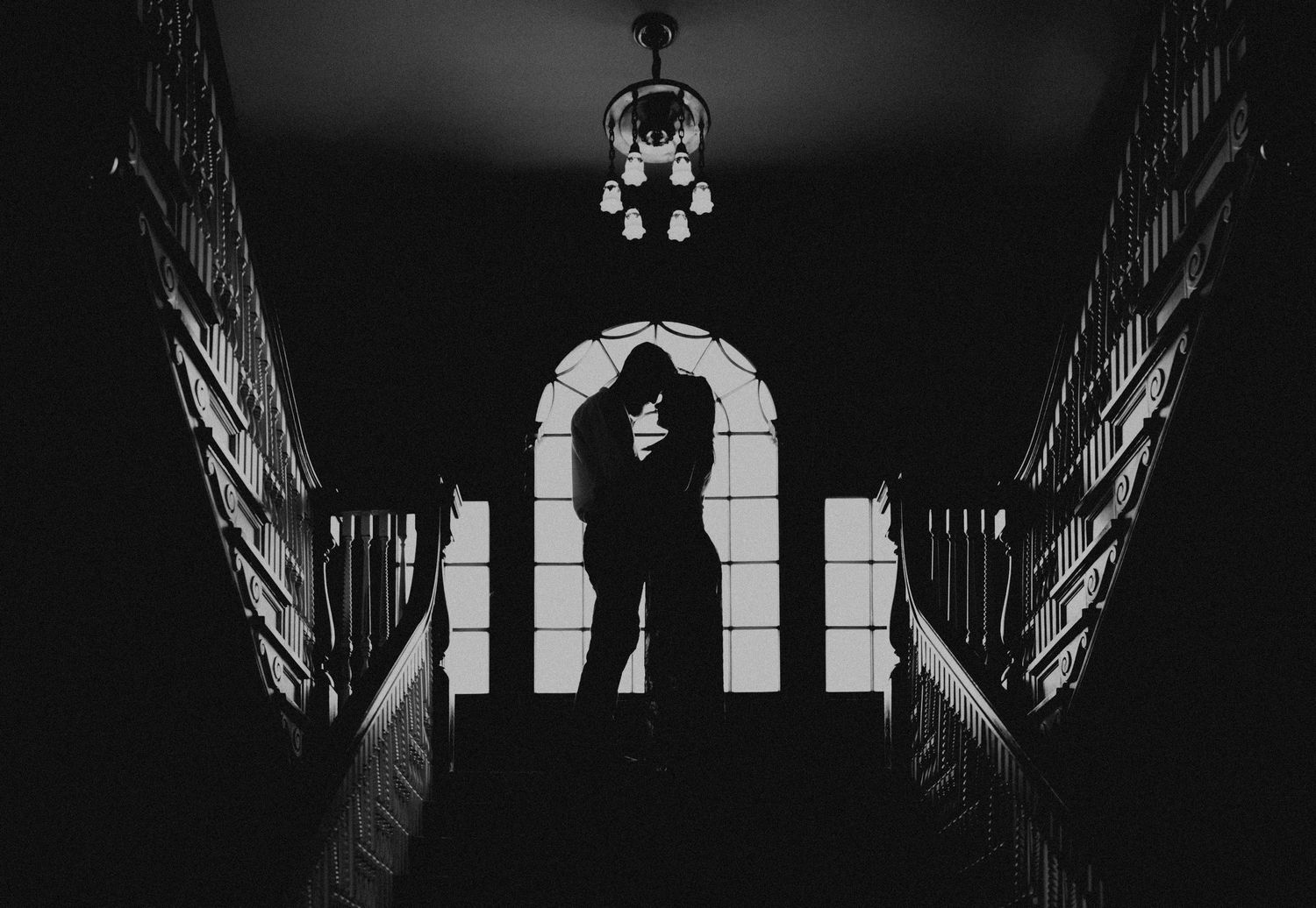 Black and white silhouette of couple embracing on staircase landing beneath elegant chandelier.