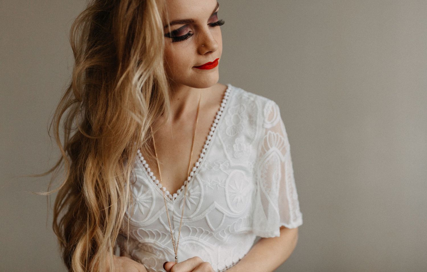 Blonde woman in white lace dress with red lips and elegant makeup, looking down gracefully.