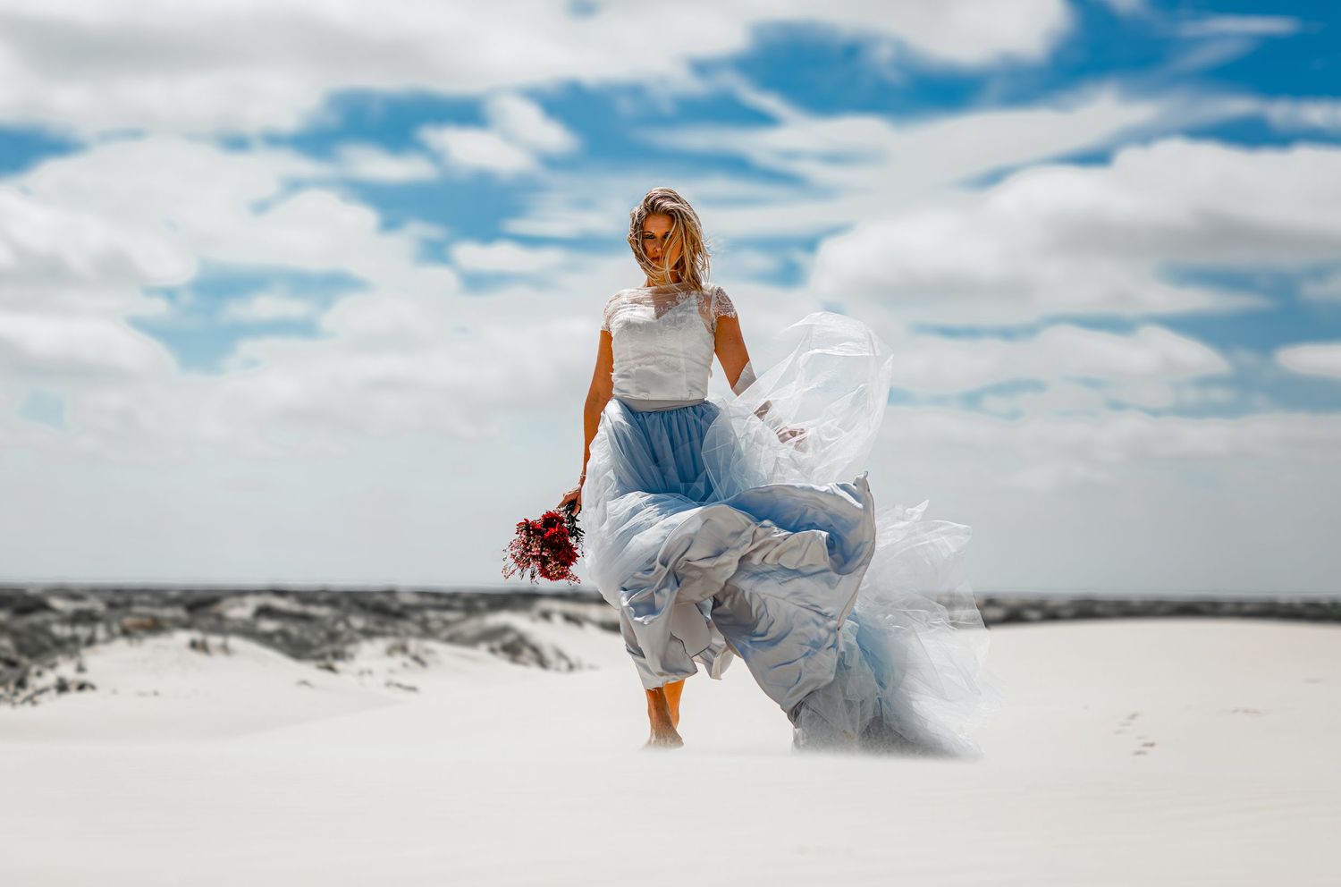 Woman in blue floral skirt holding red bouquet walks on white sand dunes under a cloudy sky.