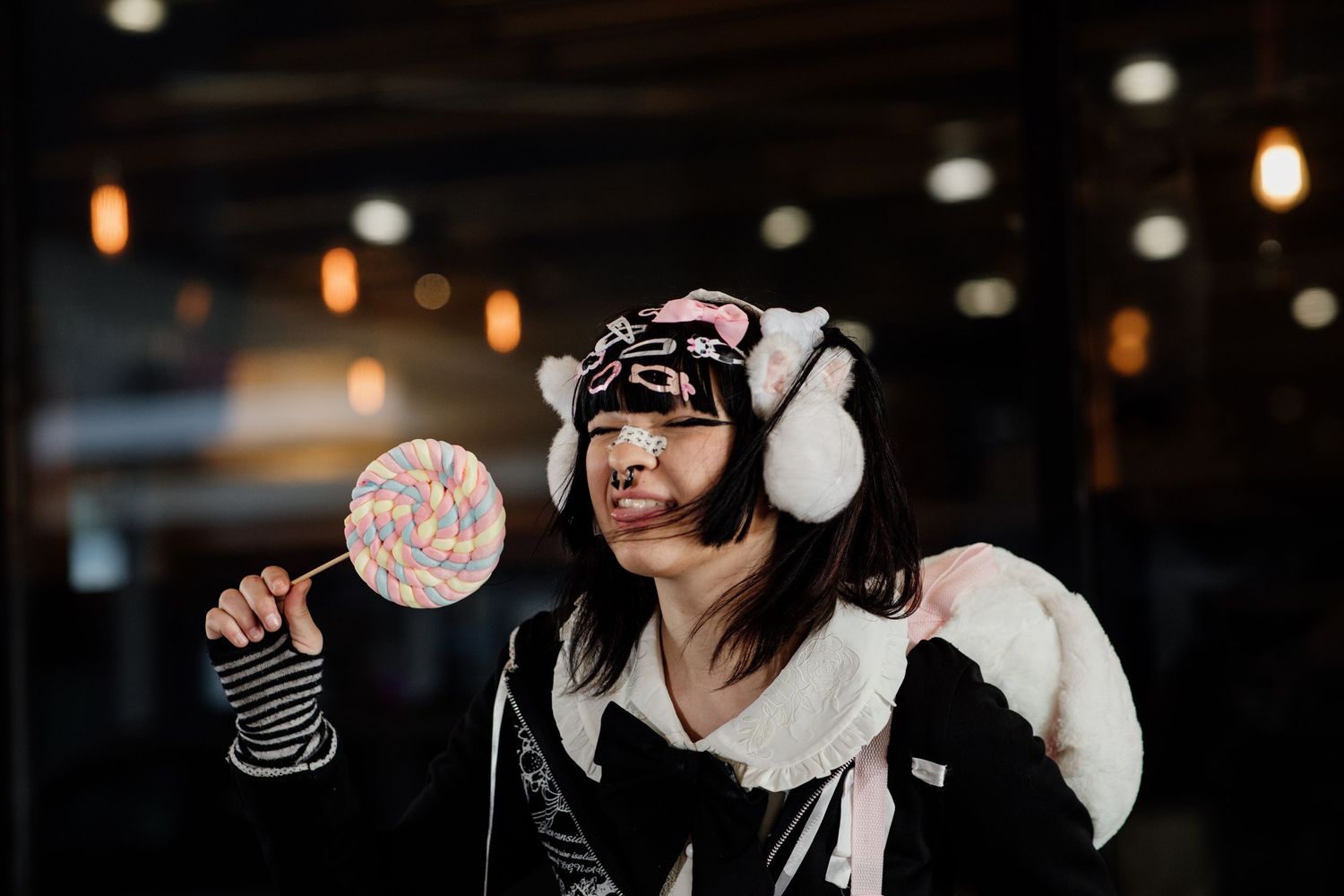Young woman in panda ear muffs and costume holds a colorful lollipop while posing dramatically indoors.