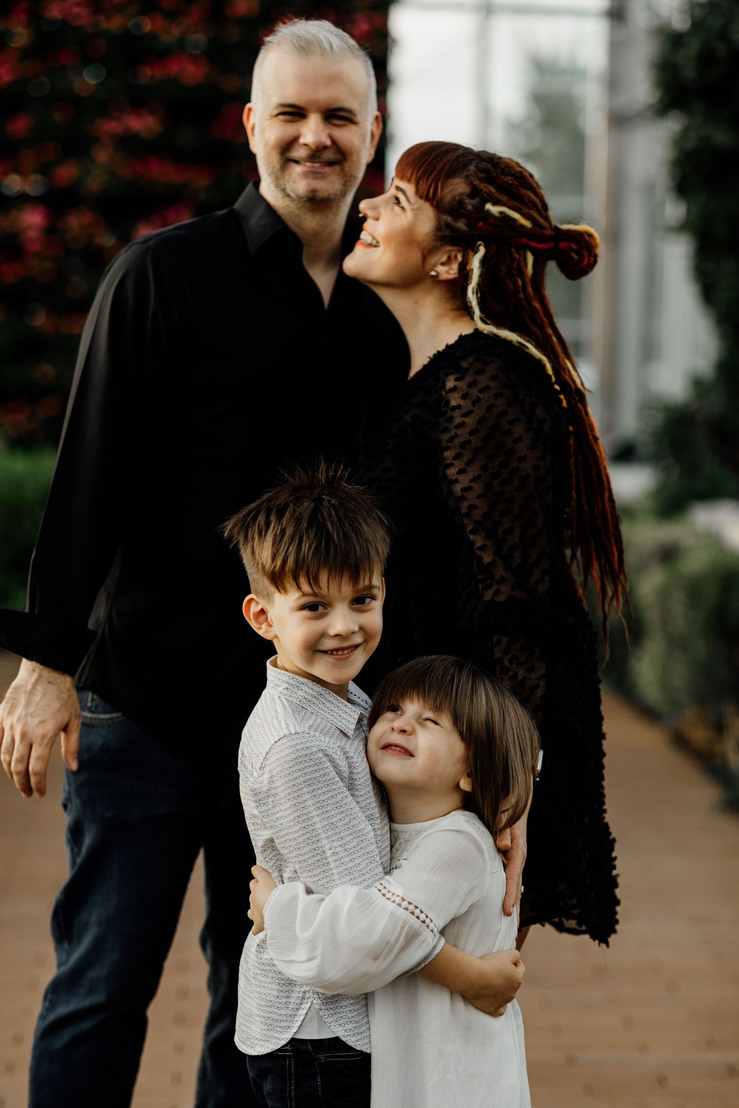 Family of four posing outdoors, parents in black attire with two young children hugging in front.