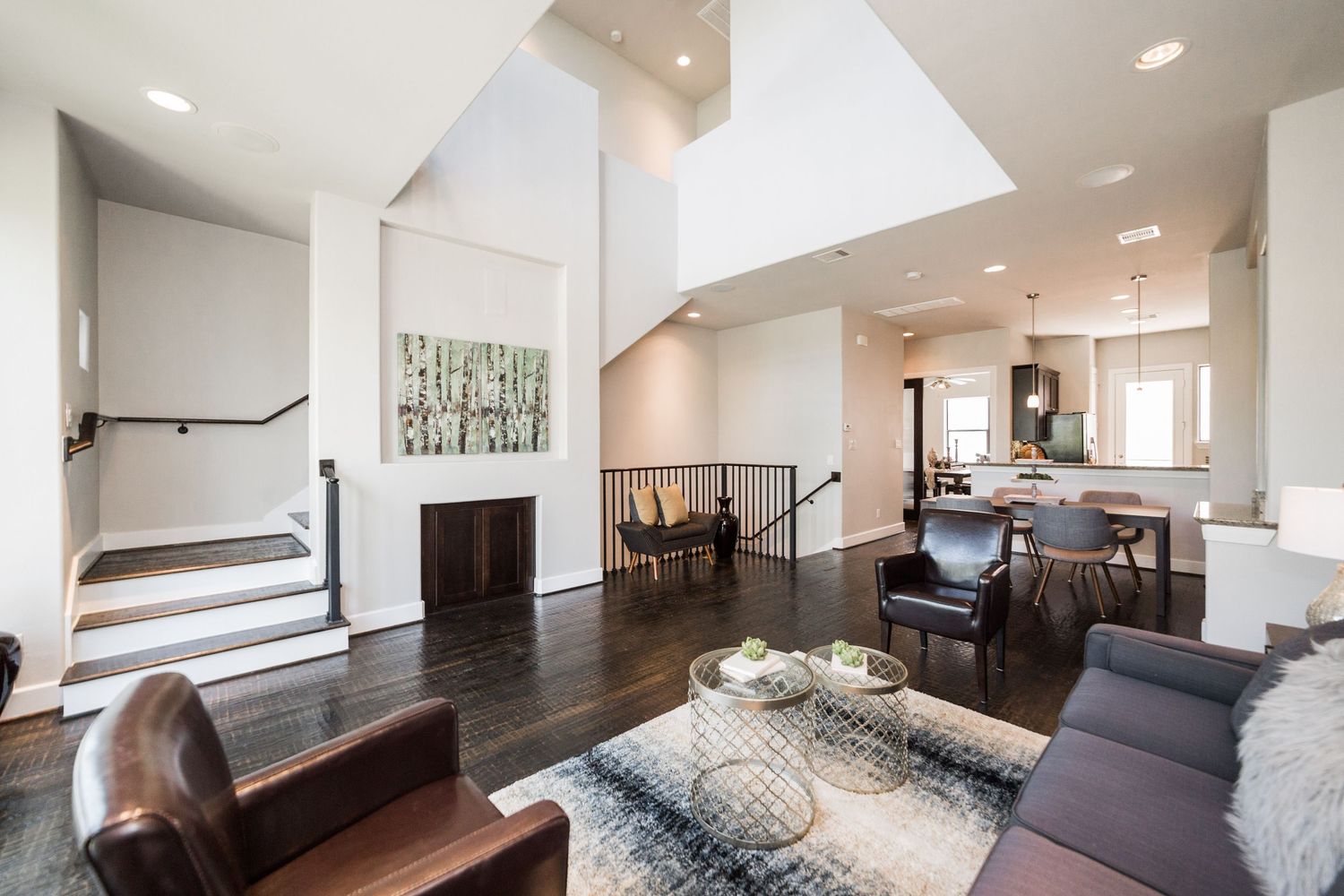 Modern open-concept living room with dark hardwood floors, high ceilings, leather chairs, and open kitchen view.
