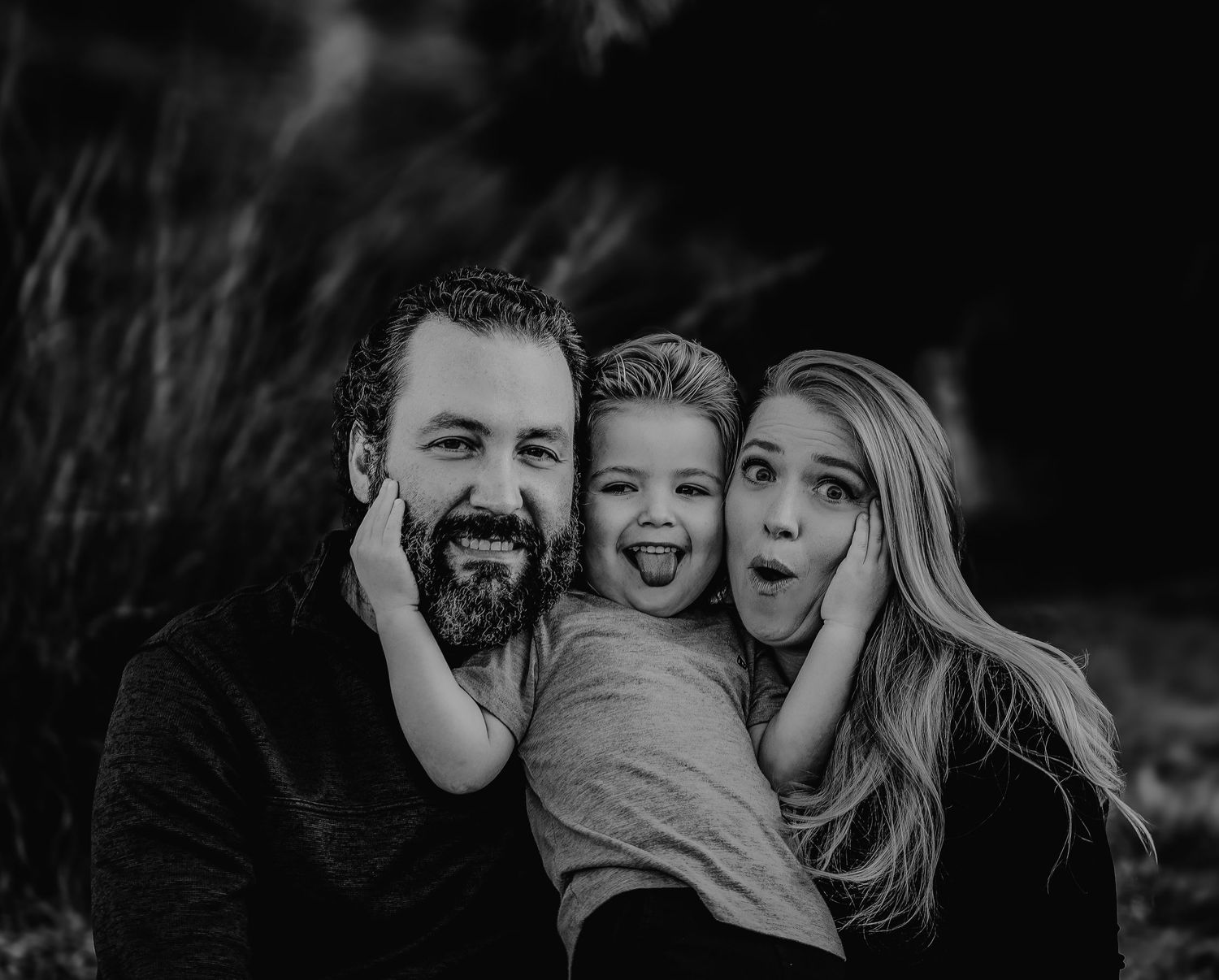Playful black and white family portrait with child sticking tongue out while parents smile outdoors.