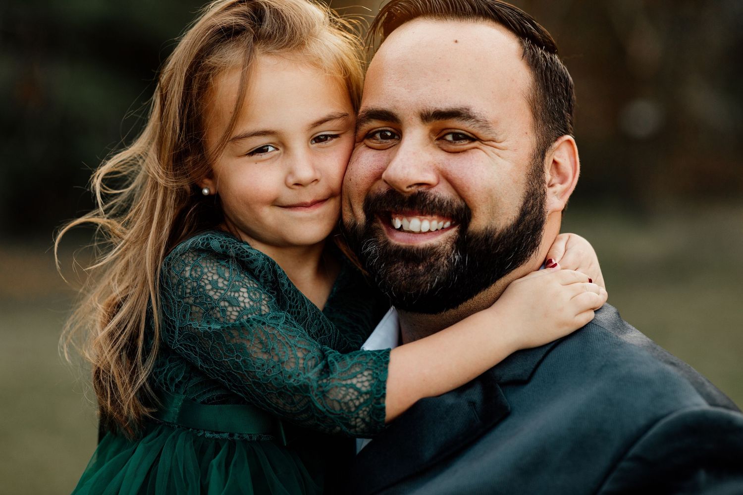 Smiling bearded father hugging his daughter in a green lace dress outdoors.