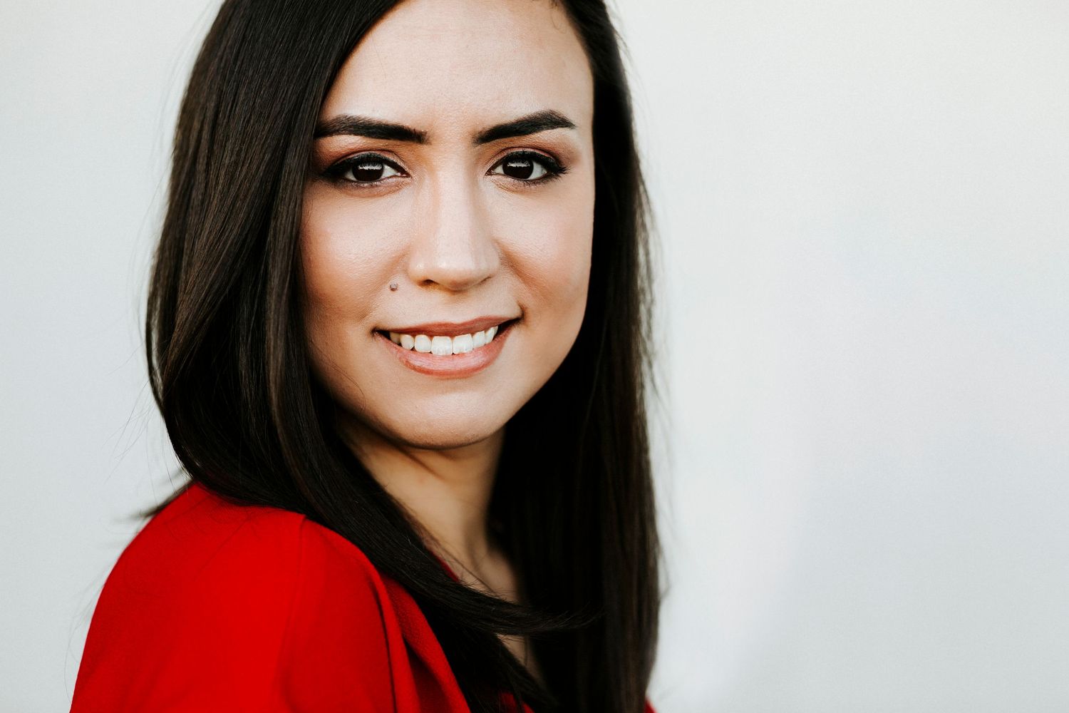 Smiling young woman with long dark hair wearing a red top against a light gray background.