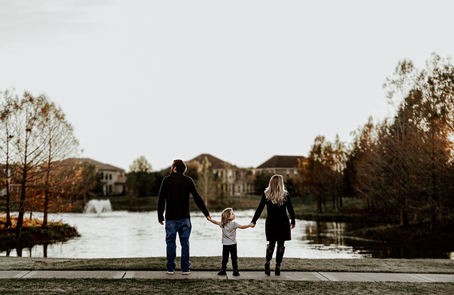 Family of three holding hands while standing near a pond with a large house in the background.