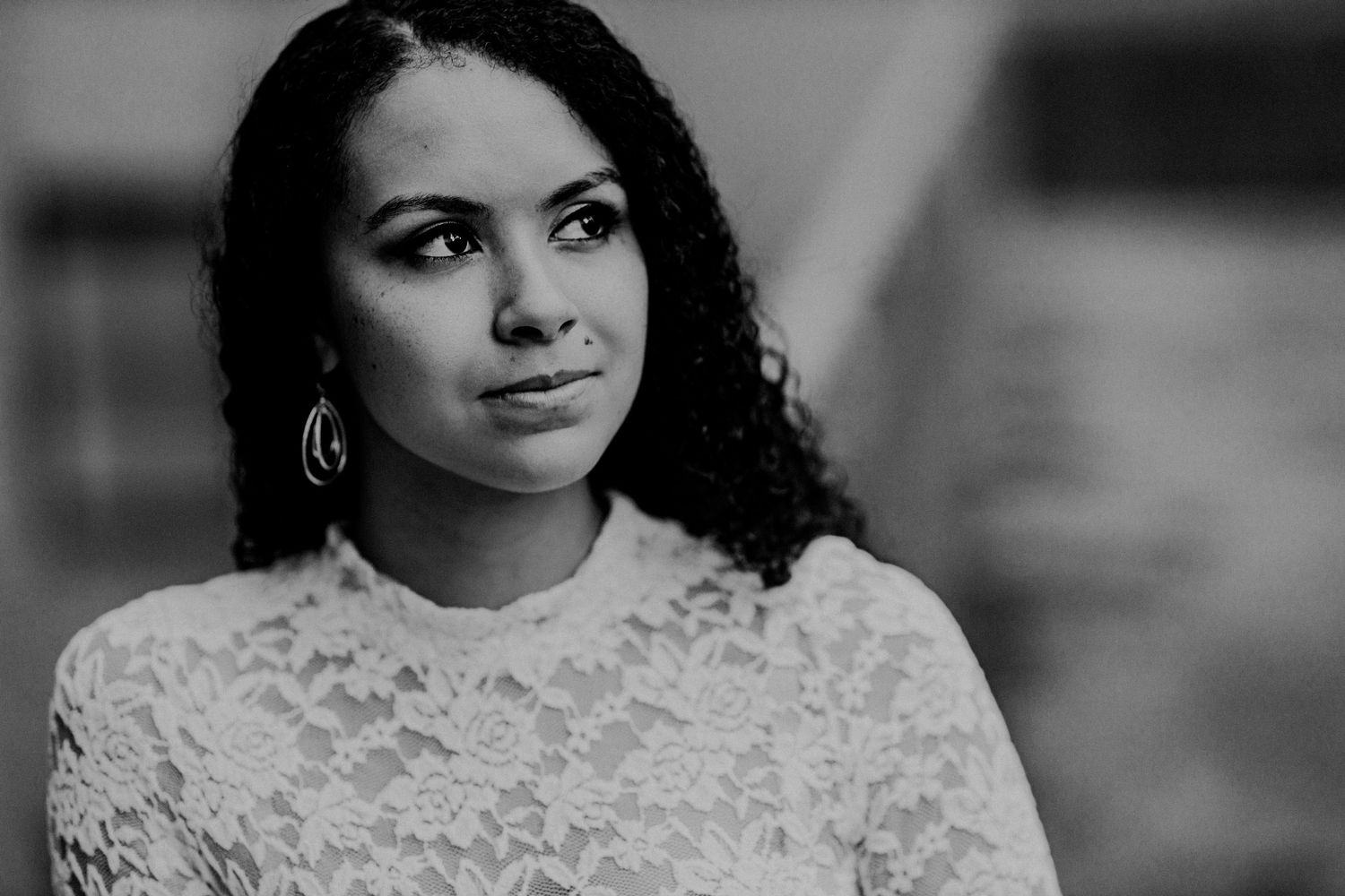 Black and white portrait of a young woman with curly hair wearing a white lace top, looking thoughtfully to the side.