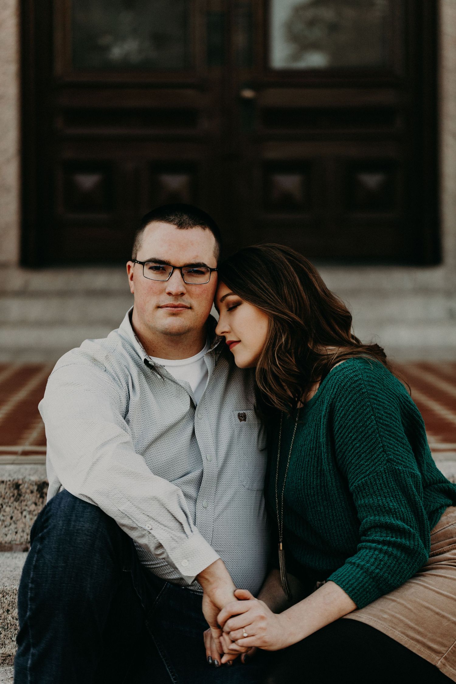 A couple sits on steps, woman in green sweater resting head on man with glasses, in front of a wooden door.
