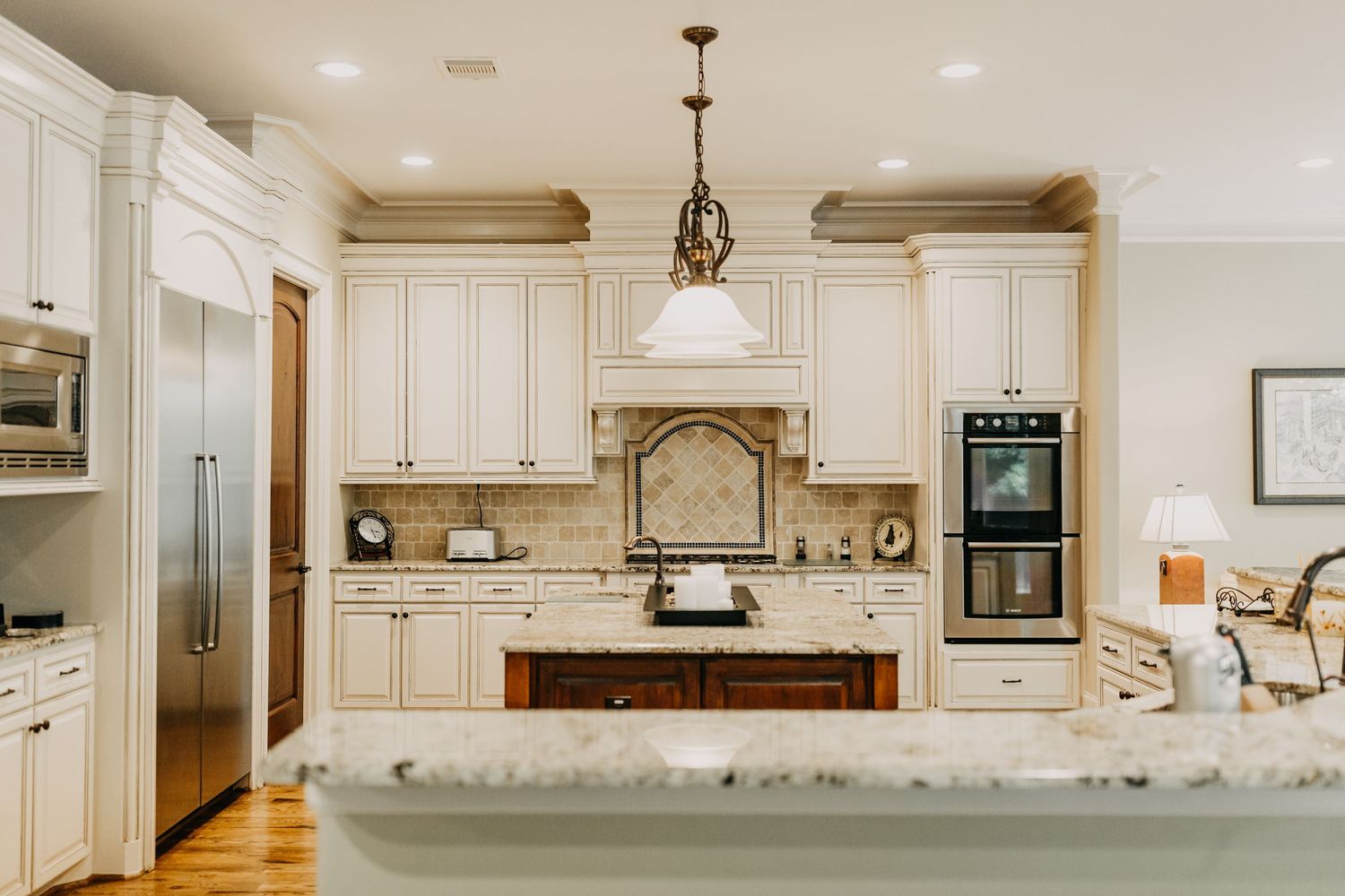 Elegant white kitchen with granite countertops, cream cabinets, stainless steel appliances, and a decorative pendant light over the island.