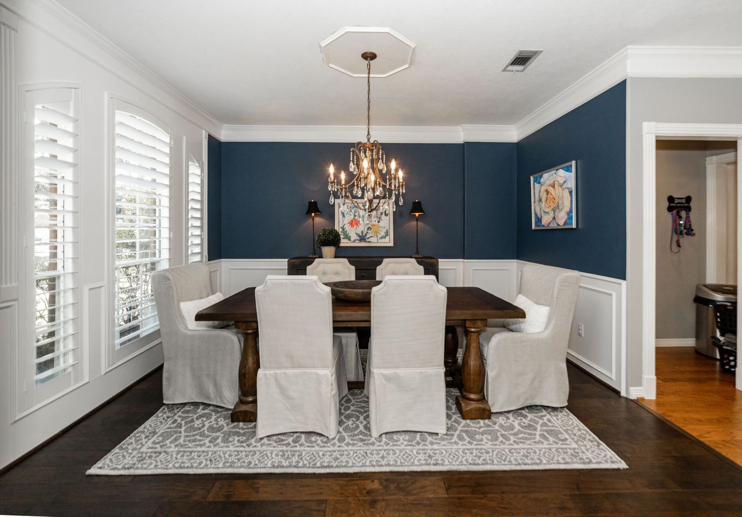 Elegant dining room with navy blue walls, white wainscoting, dark wood table, slipcovered chairs, and a black chandelier.