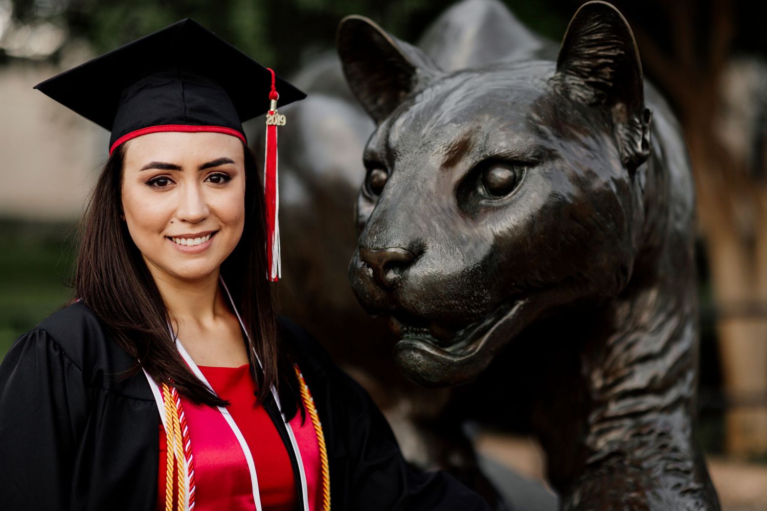 Female graduate in cap and gown smiling beside a bronze wildcat statue at university graduation.