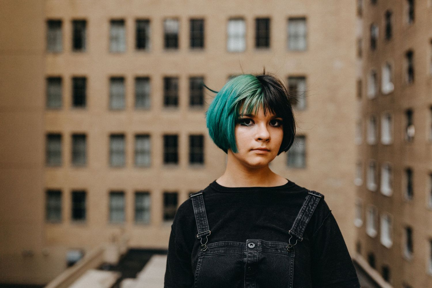 Young woman with teal bob haircut wearing black overalls stands on urban rooftop with brick building backdrop.