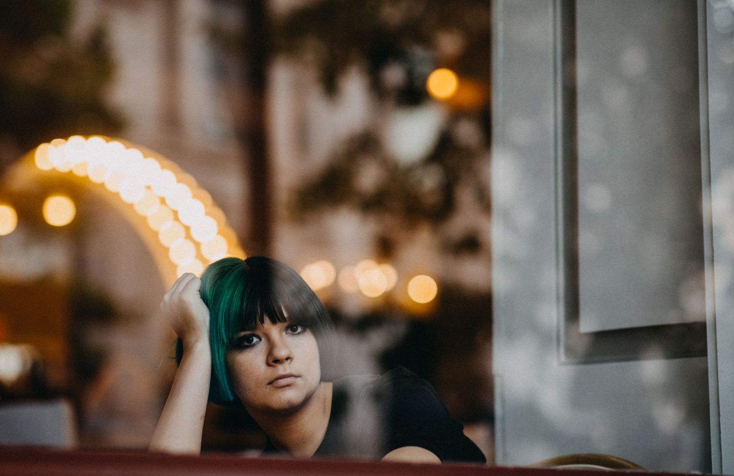 Young woman with teal green hair gazing thoughtfully through a cafe window with bokeh lights in background.