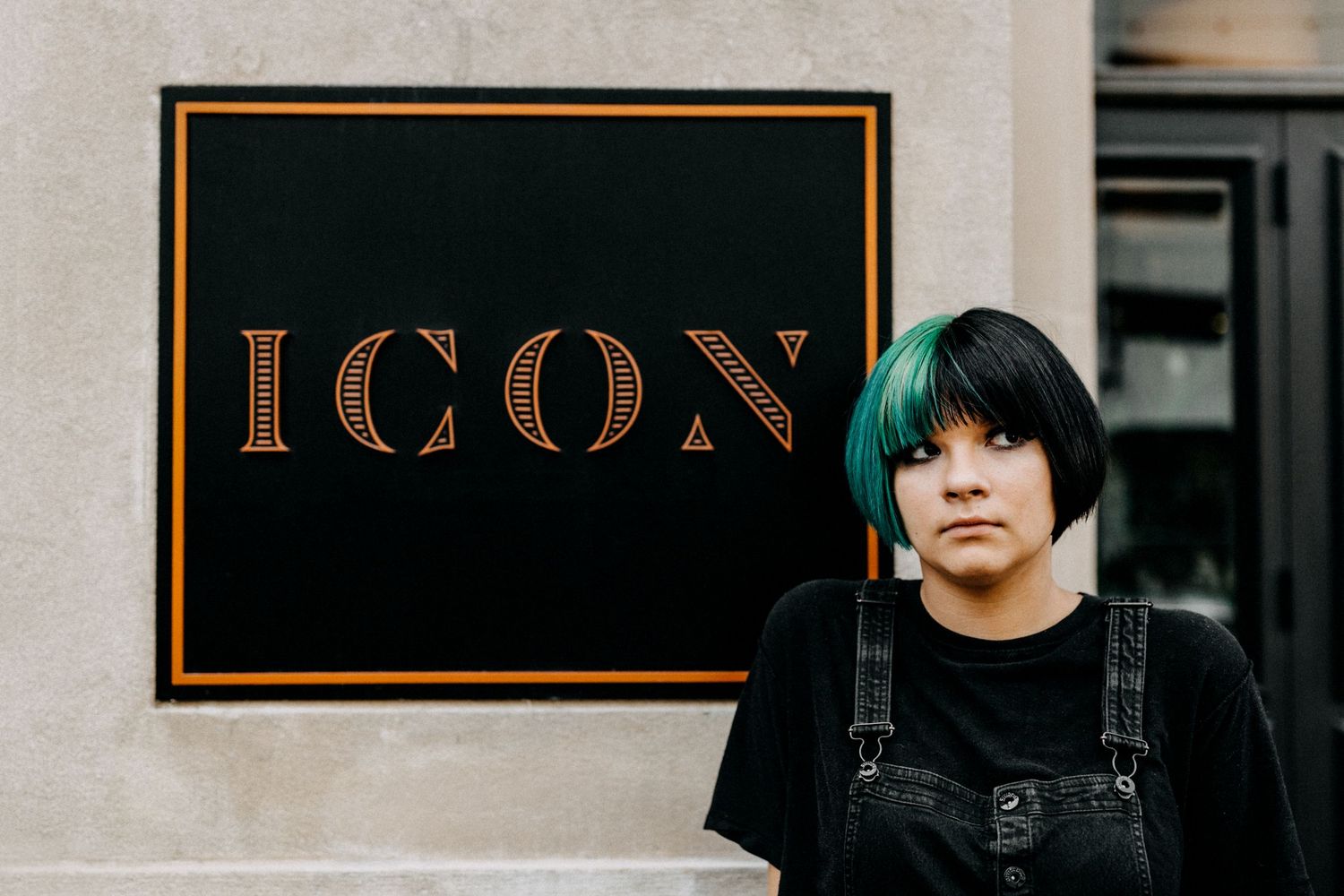 Young woman with black and green hair standing in front of an ICON sign wearing black overalls outdoors.