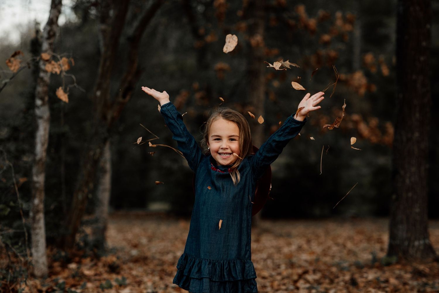 A joyful young girl in a teal dress tosses autumn leaves in the air while smiling in a fall forest setting.