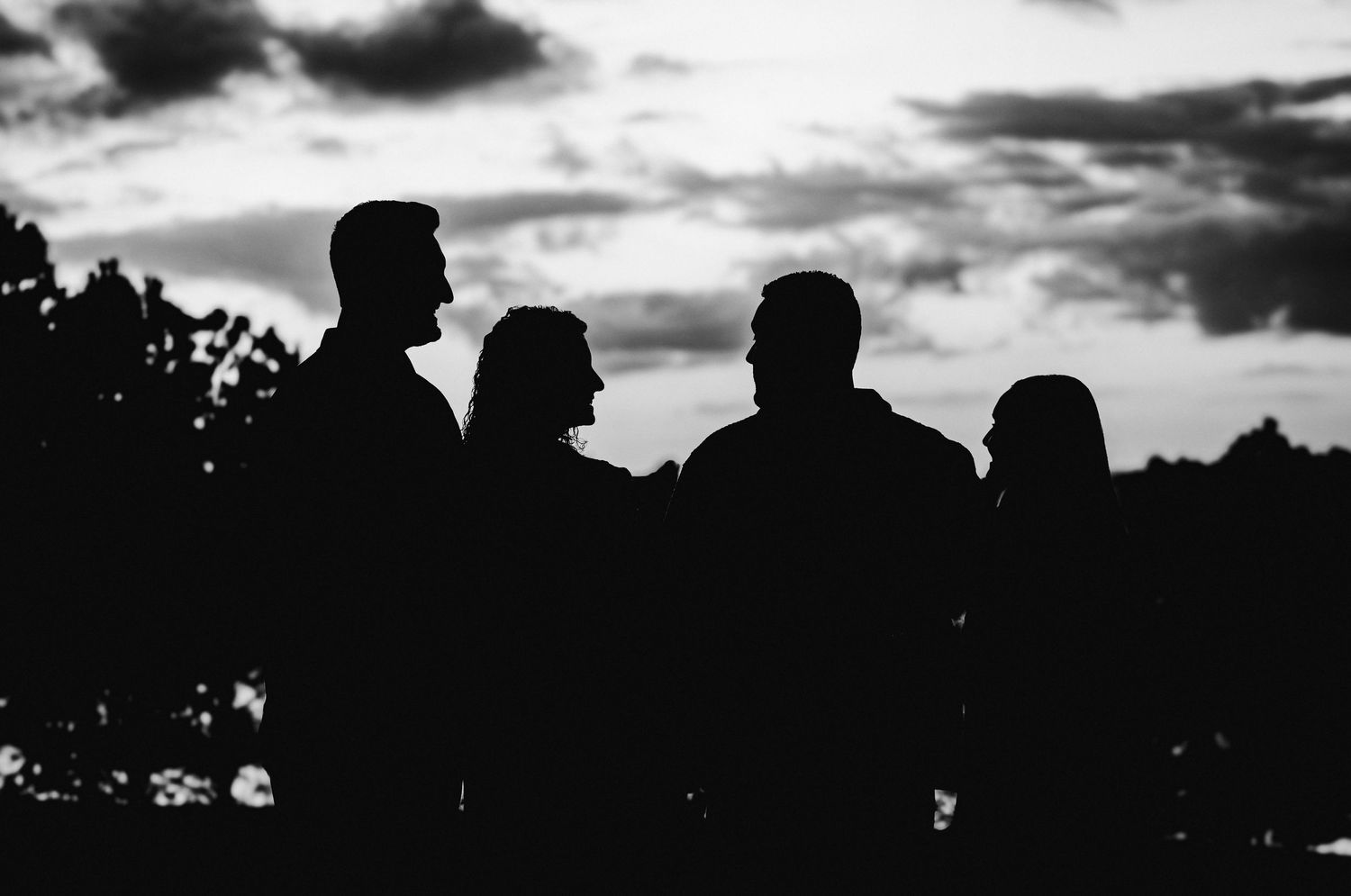 Black and white silhouette of four people standing outdoors against a dramatic cloudy sky at dusk.
