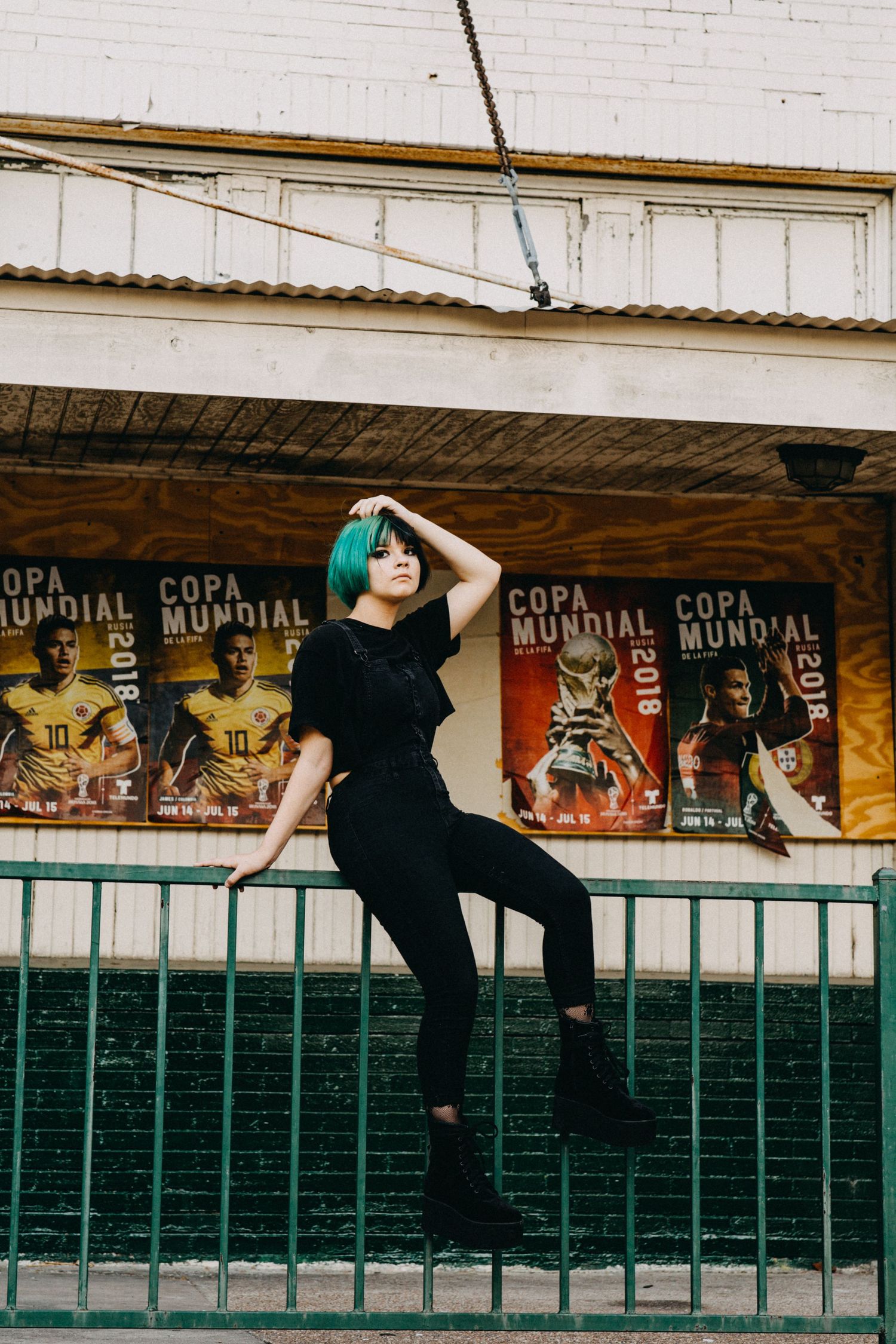 Woman with teal hair in black outfit posing against green fence with Copa Mundial 2018 soccer posters.
