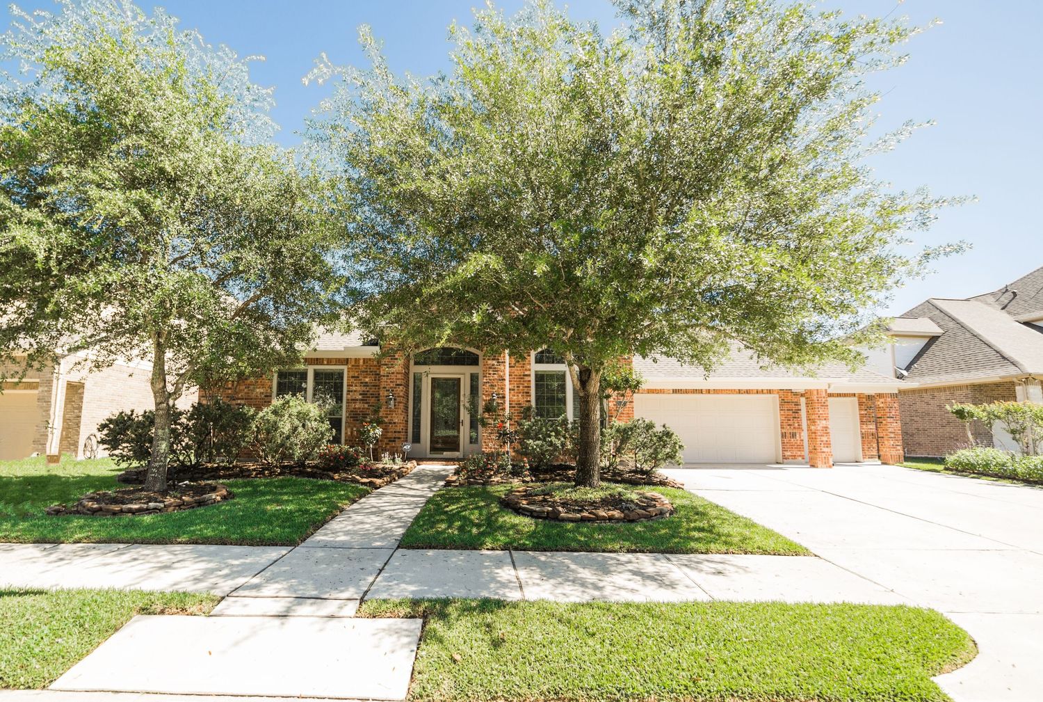 Brick suburban home with large shade tree, three-car garage, manicured lawn, and landscaped front yard on a sunny day.
