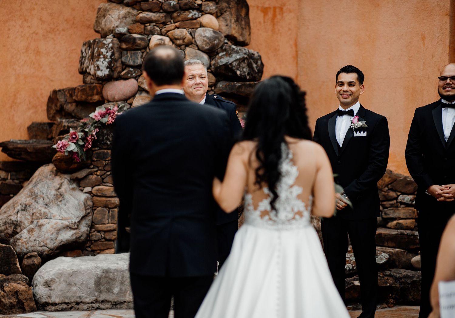 Bride and groom stand before smiling officiant during outdoor wedding ceremony with stone wall backdrop.