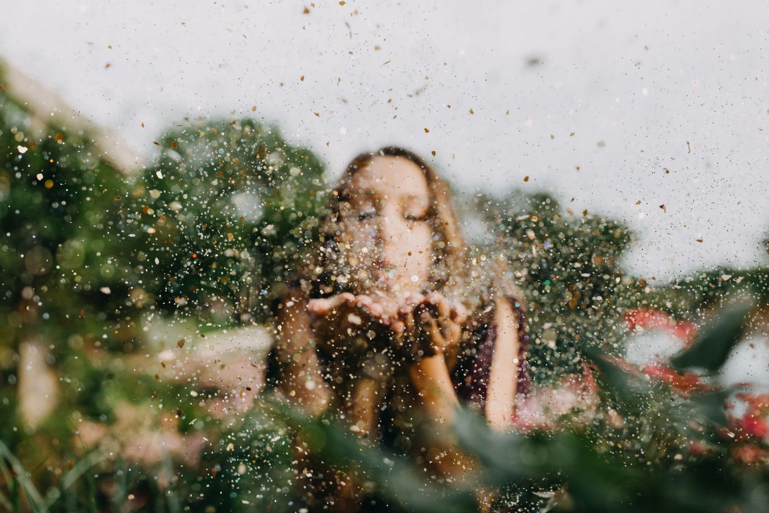 A young woman blowing golden glitter from her hands outdoors, surrounded by lush green foliage.