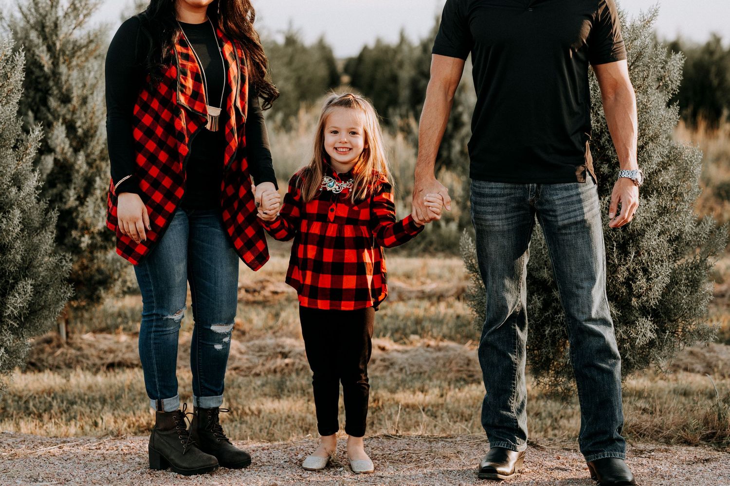 Family of three in matching red plaid shirts holding hands at a Christmas tree farm.