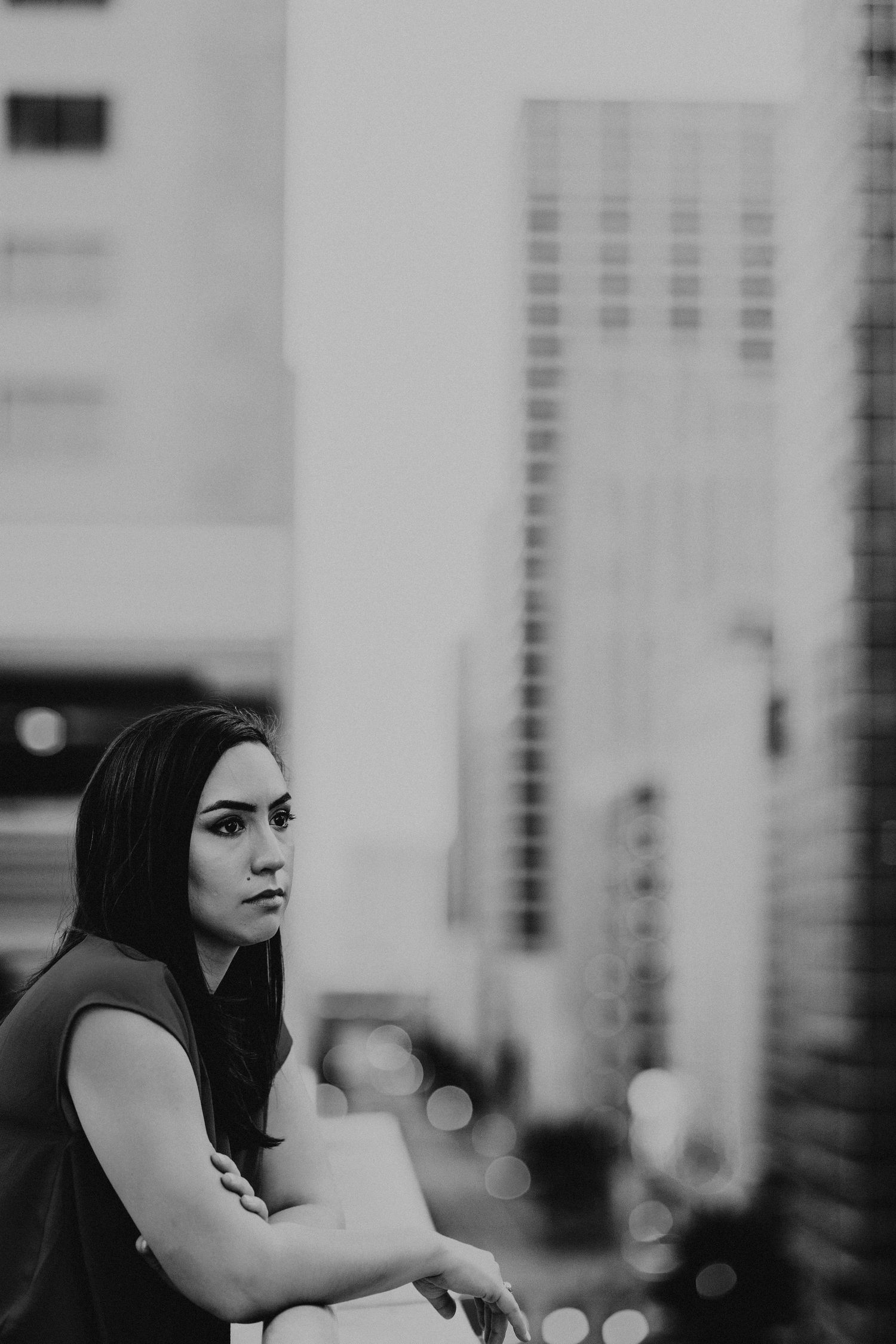 Black and white portrait of a young woman with long hair standing in an urban cityscape with skyscrapers.