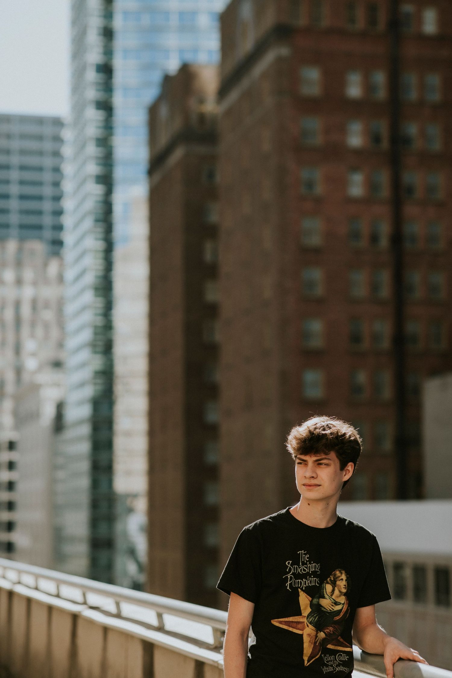 A young man in a graphic tee stands on a urban rooftop with city buildings behind him.