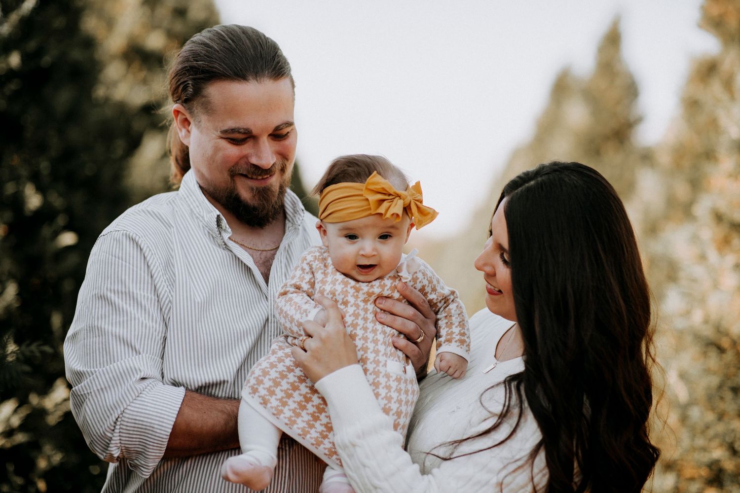 Happy young family with parents smiling and holding their baby girl in a mustard yellow bow outdoors.