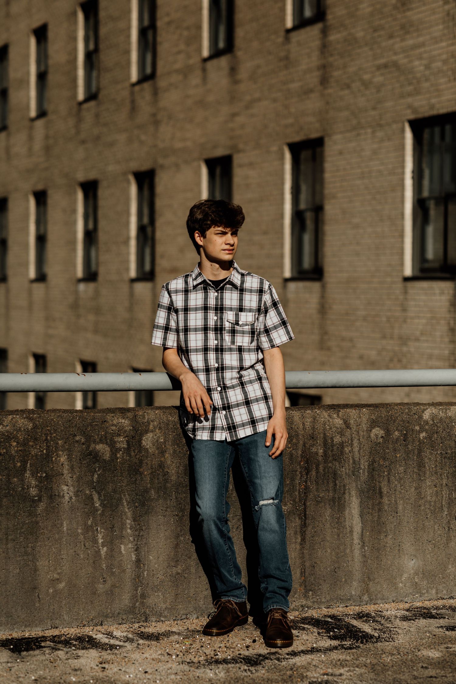 Young man in plaid shirt and jeans leaning against concrete wall with brick building backdrop in urban setting.