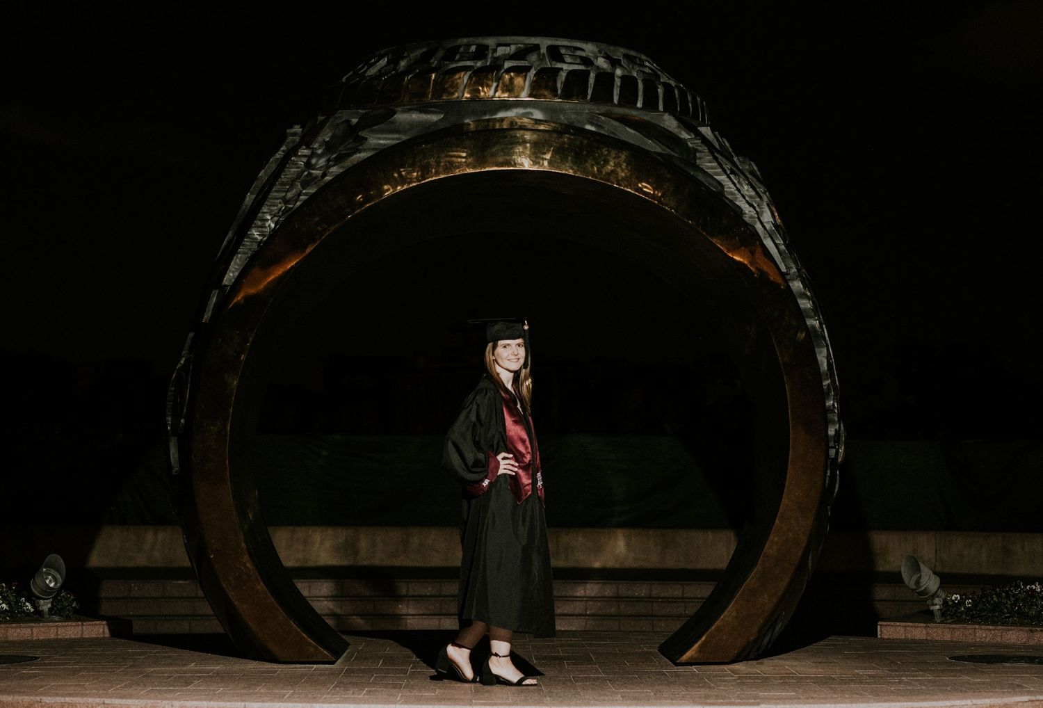Graduate in cap and gown poses before a large decorative ring sculpture at night.