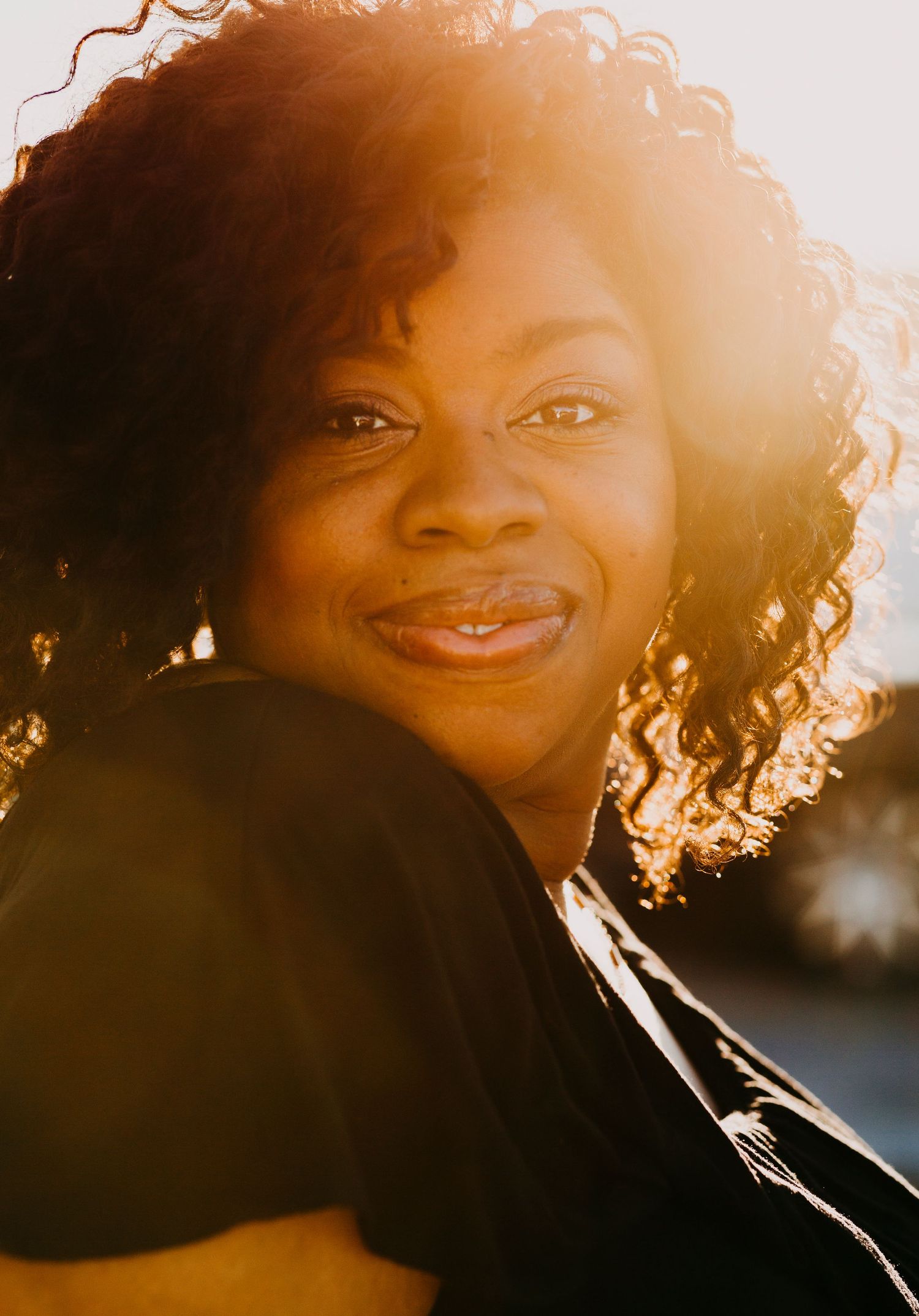 A smiling Black woman with curly hair bathed in golden sunset backlight outdoors.