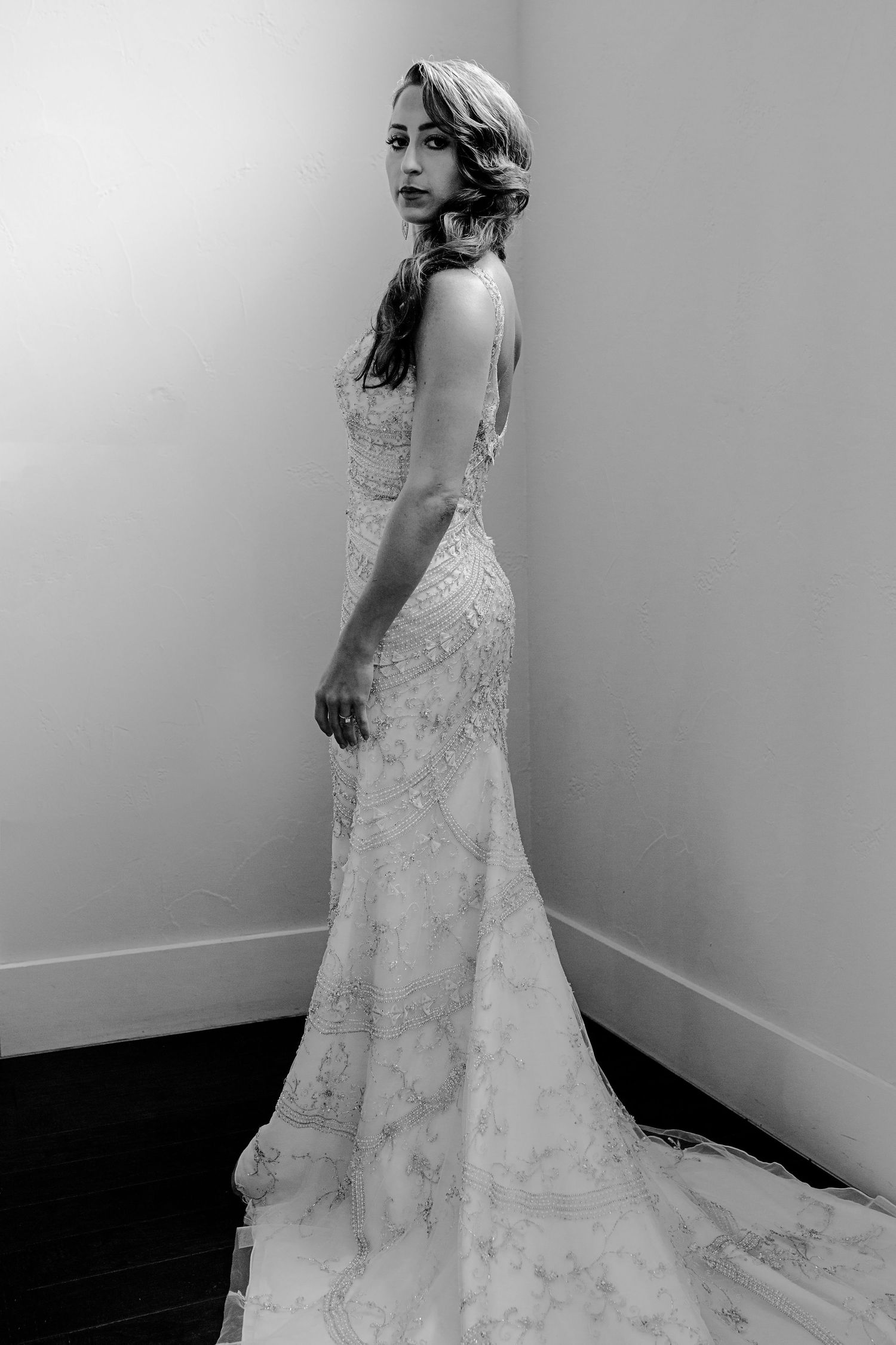Black and white photo of a bride in an elegant lace wedding gown with a train, posing gracefully in a corner.