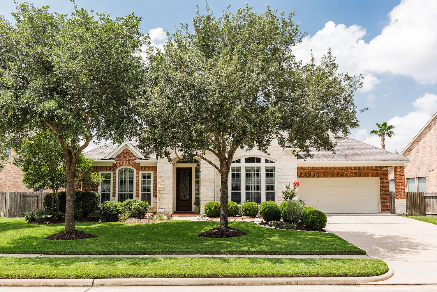 Single-story brick home with large shade tree, manicured lawn, two-car garage, and arched windows on a sunny day.