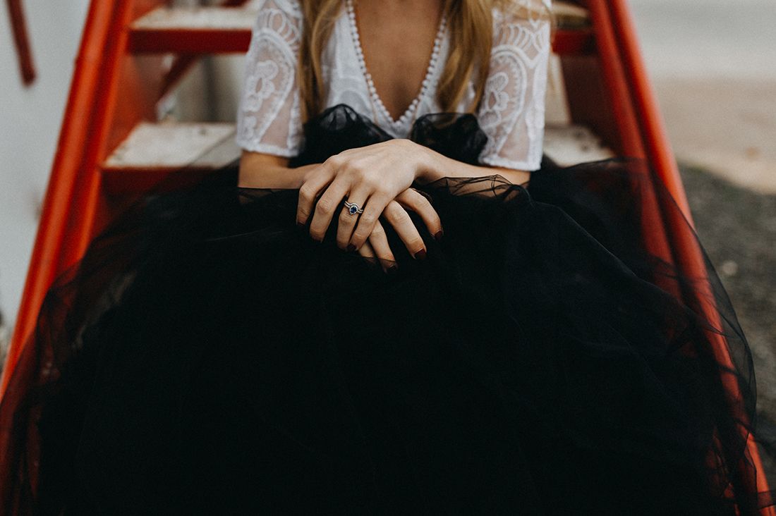 Bride in white lace top and black tulle skirt with hands clasped on red chair.