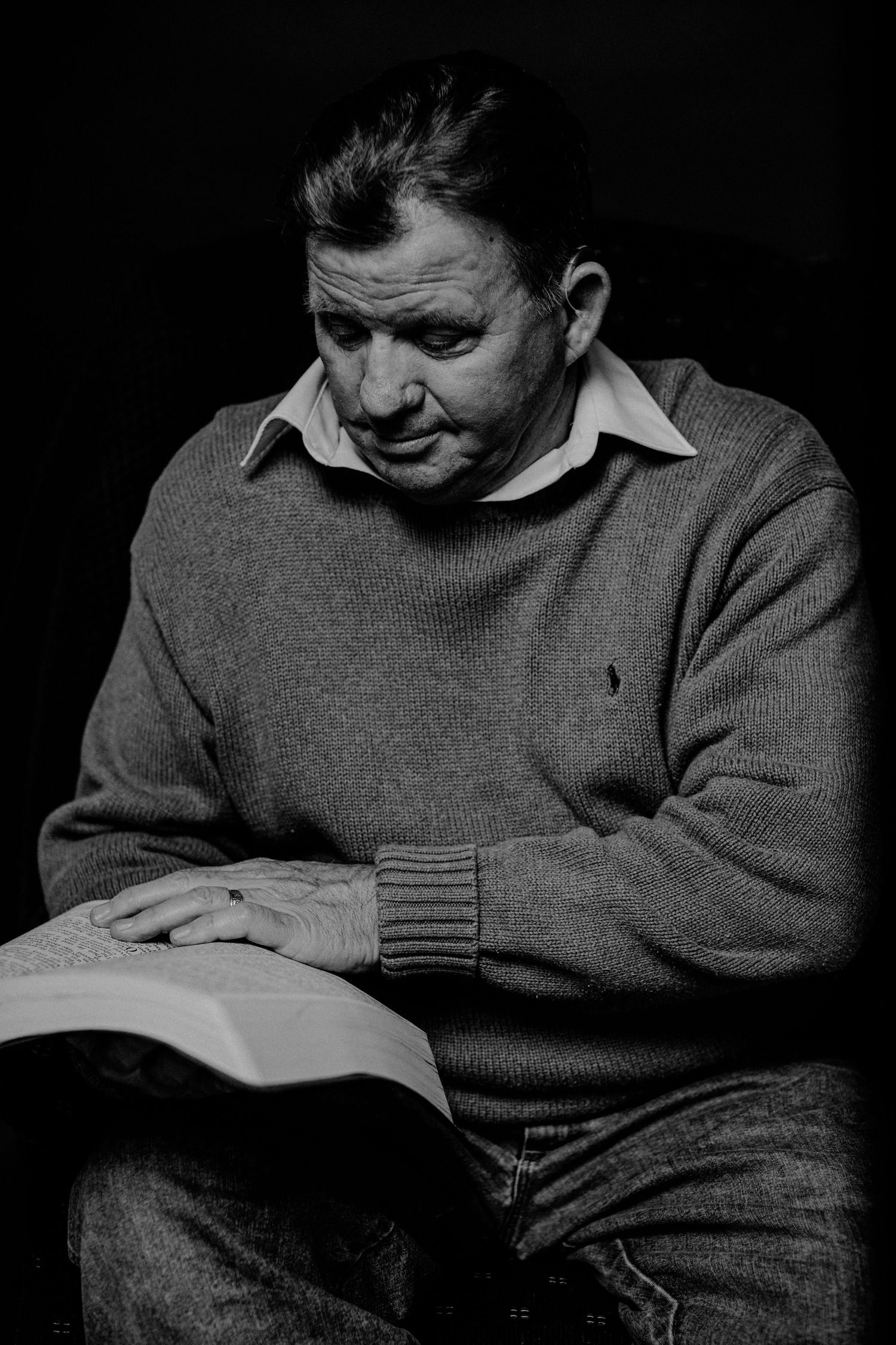 Black and white portrait of a middle-aged man in a sweater reading a book against a dark background.