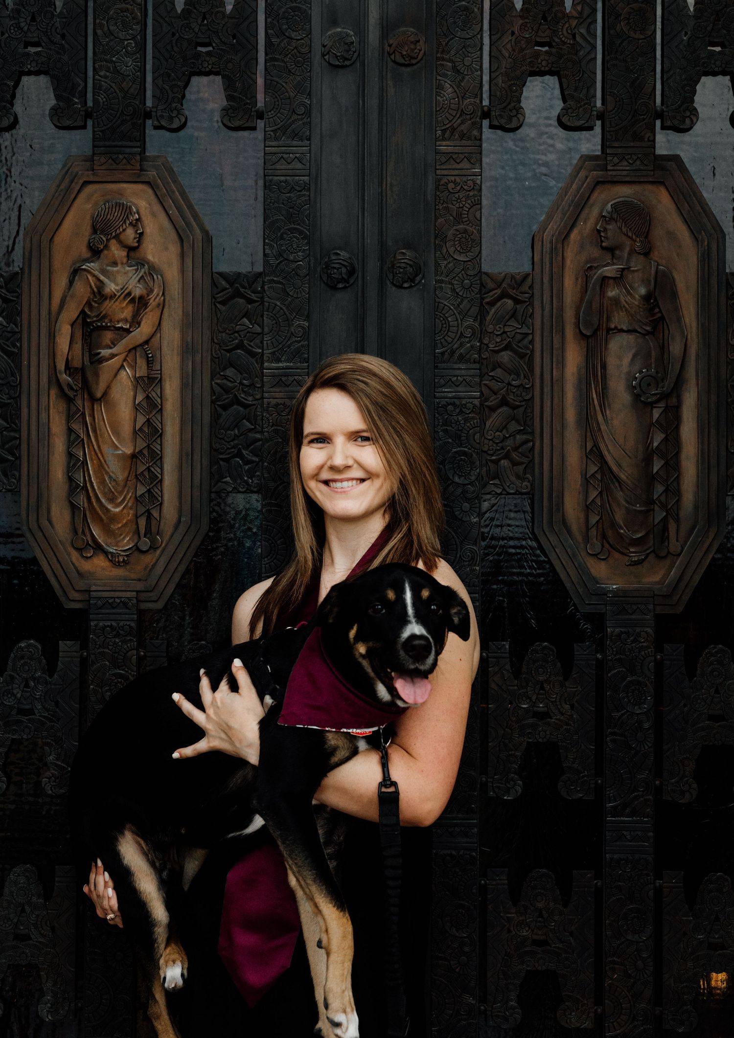 Smiling woman holding a black and white dog in front of ornate bronze doors with classical relief sculptures.