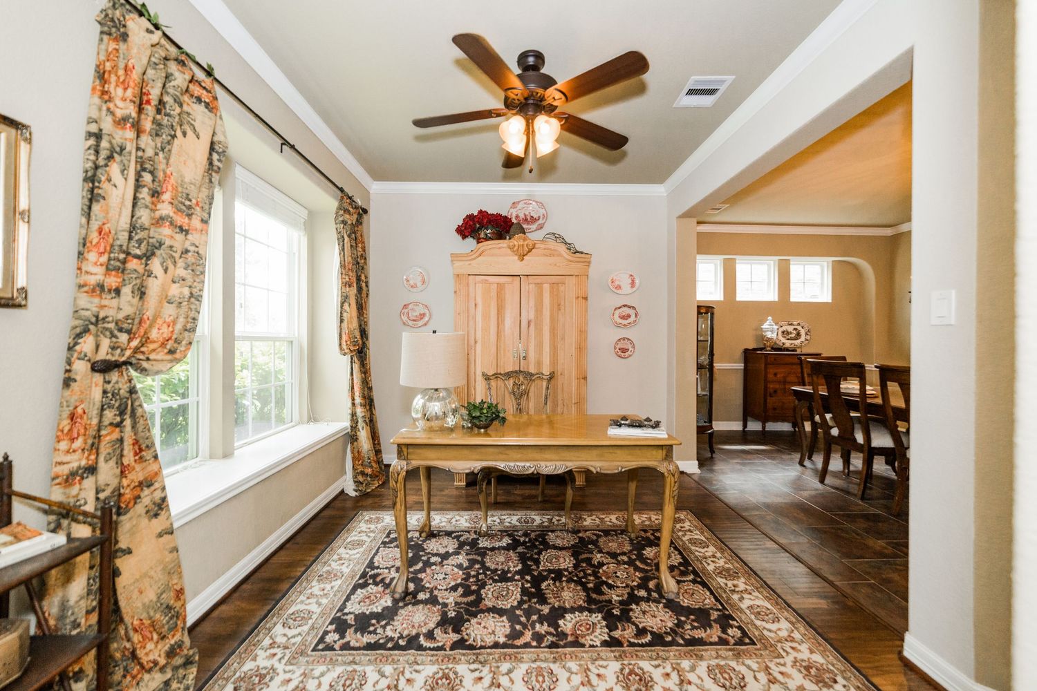 Elegant dining room with ornate rug, antique wooden desk, large armoire, floral curtains, and ceiling fan on hardwood floors.