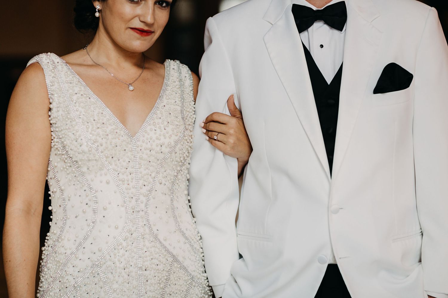 Bride in pearl-embellished white dress standing beside groom in white tuxedo.