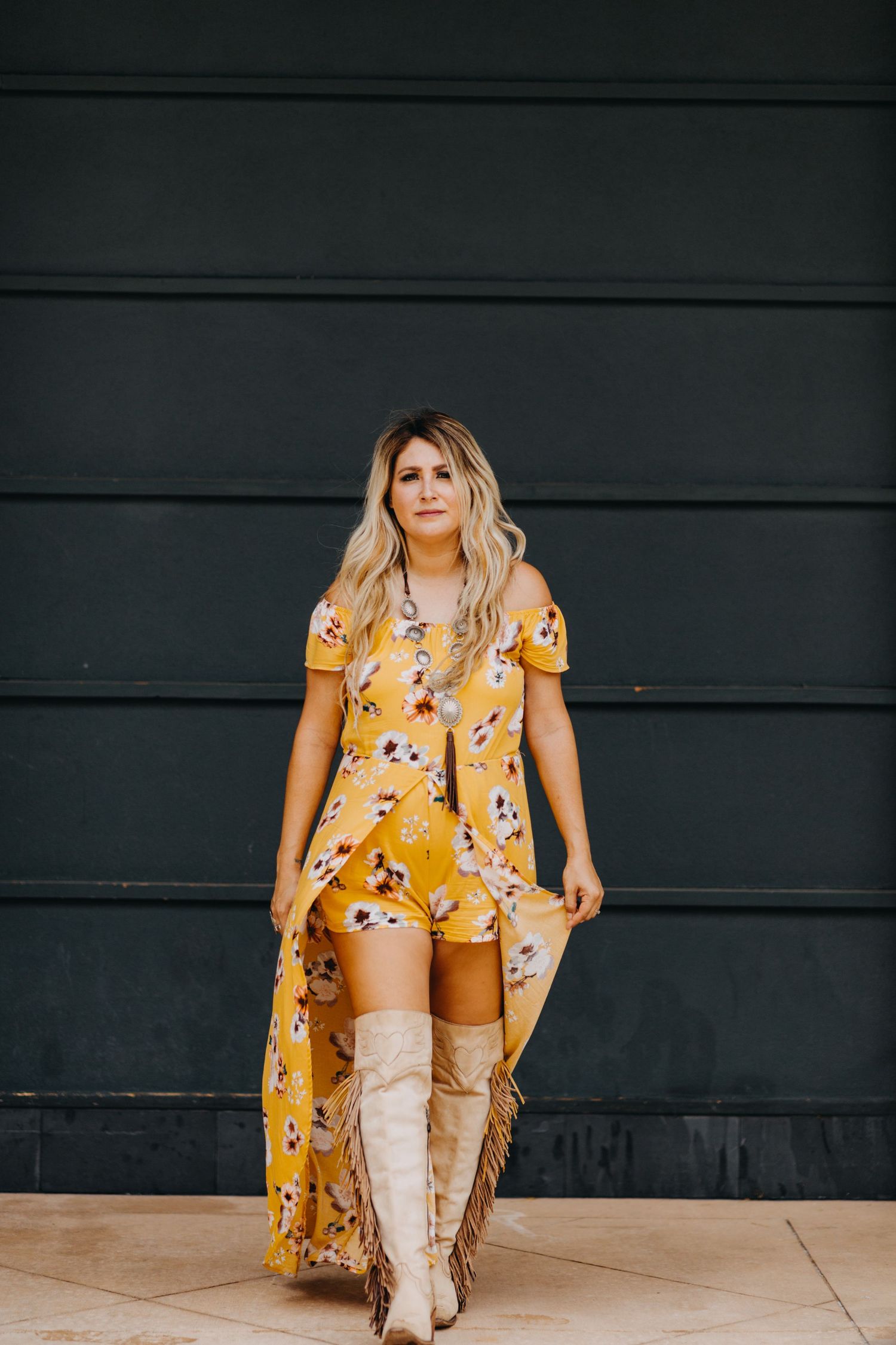 A woman in a yellow floral maxi dress and fringe boots poses against a dark green wall.