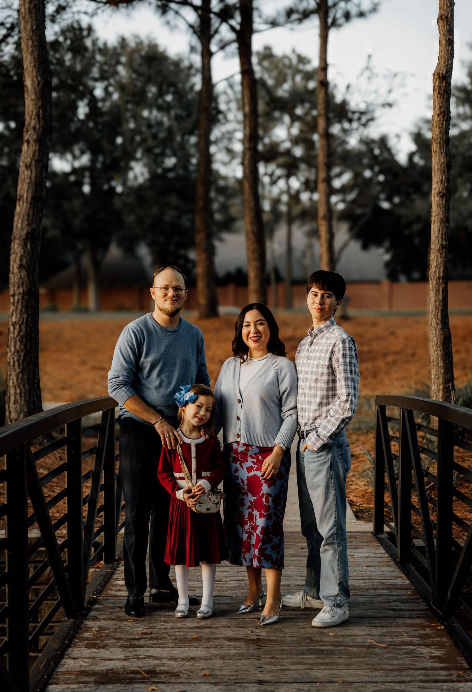 Family of four posing on a wooden bridge surrounded by tall pine trees during an outdoor portrait session.