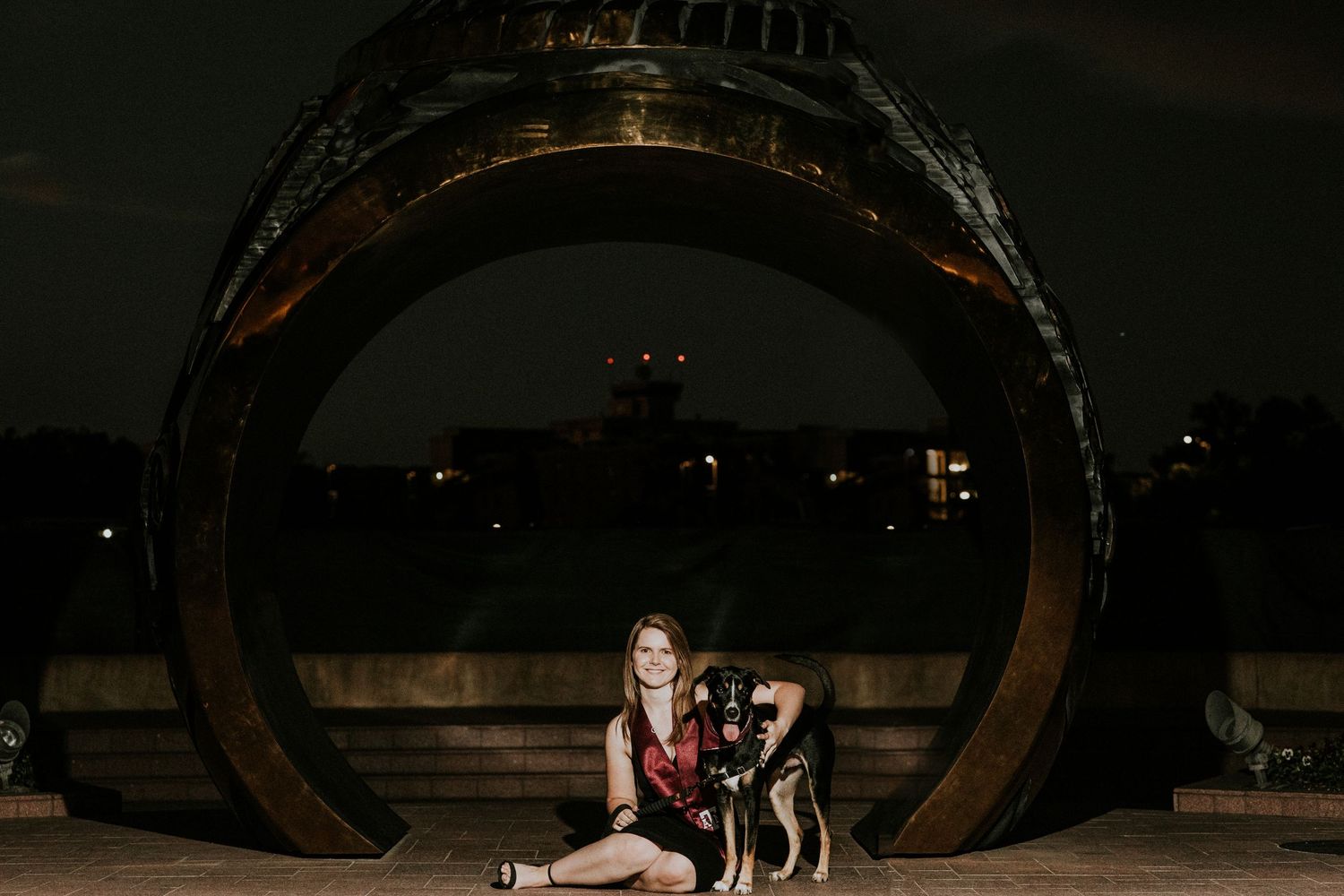 Woman sitting with dog in front of large ring sculpture at night.