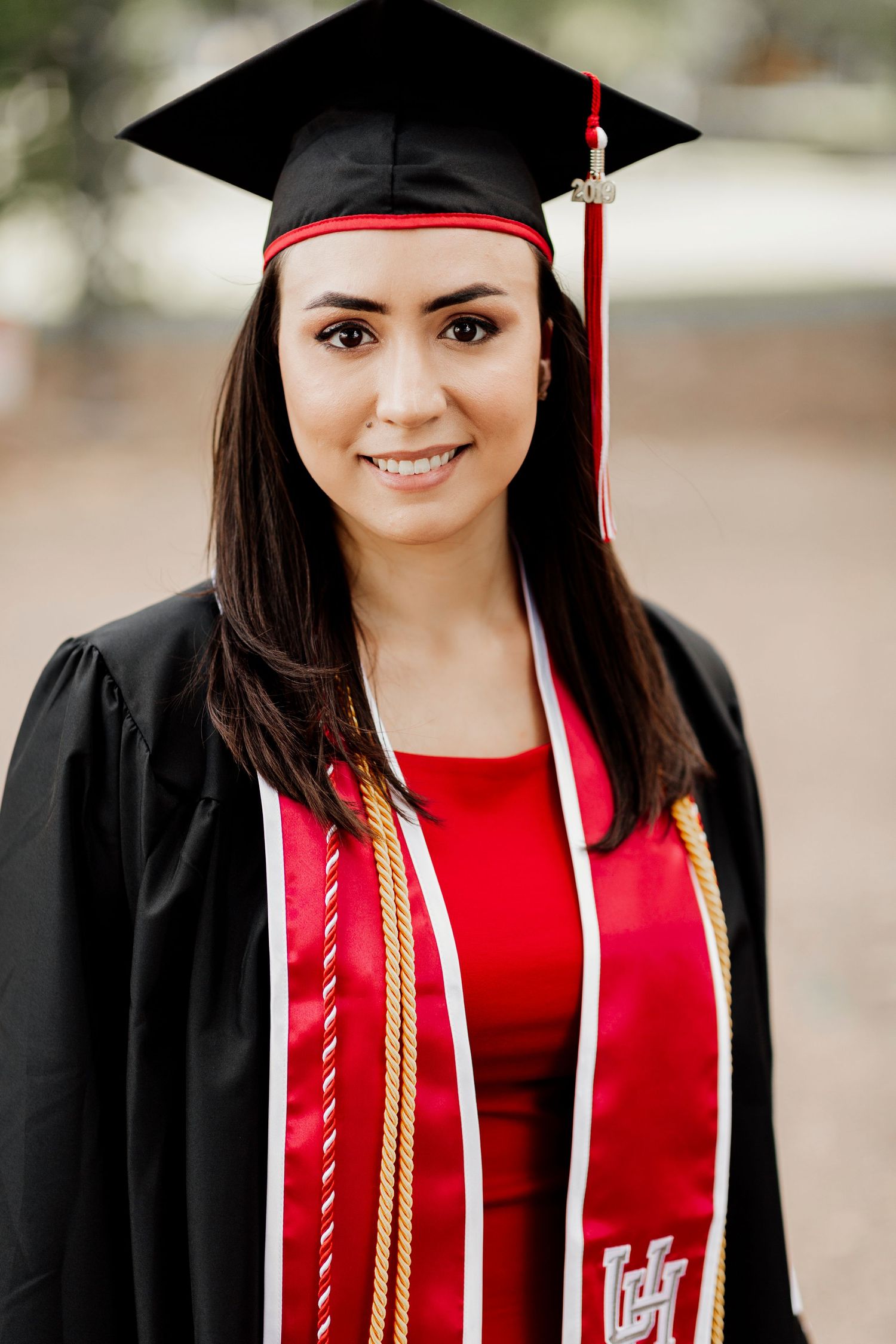 Young woman in black graduation gown and cap with red UH stole and honor cords smiling outdoors.