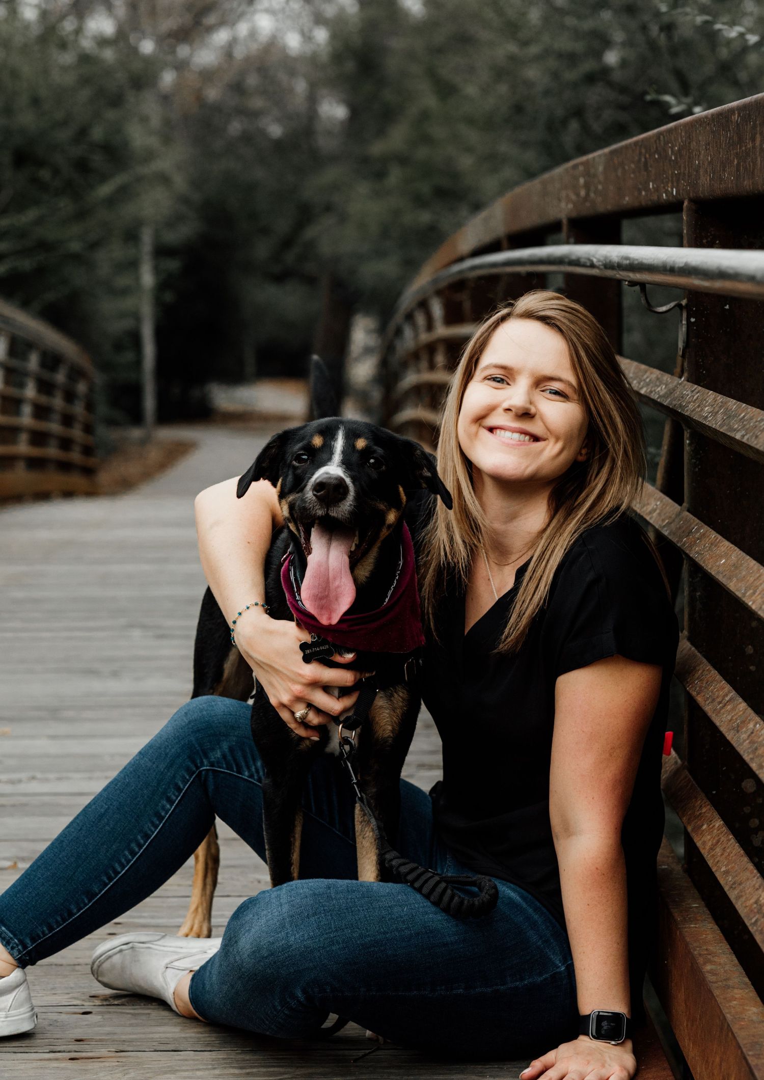 Smiling woman sitting on a wooden bridge with a black and tan dog, both posing outdoors in a park setting.