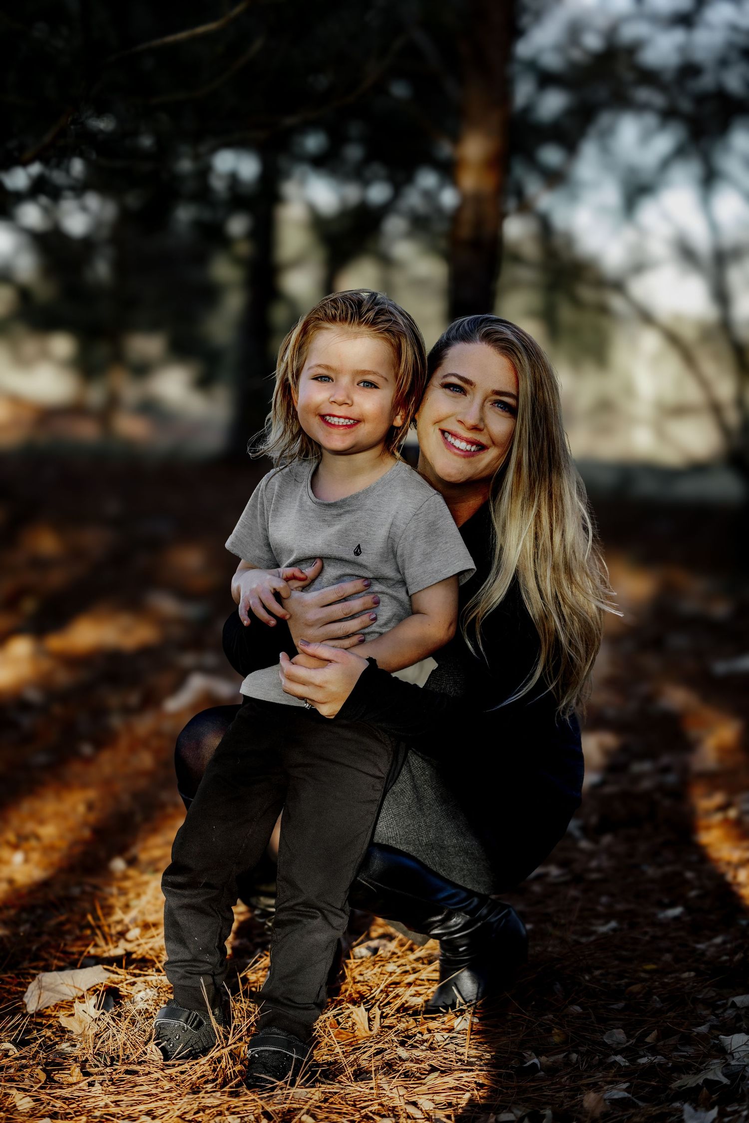 A smiling woman in black crouches beside a cheerful young child sitting on a rock in autumn woods.