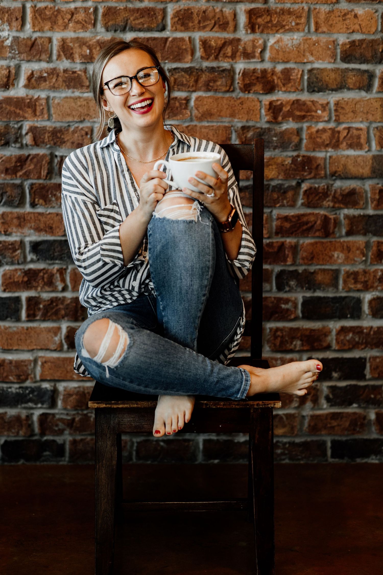 Smiling woman in glasses, striped shirt, and ripped jeans sitting on a chair holding a coffee mug against a brick wall.
