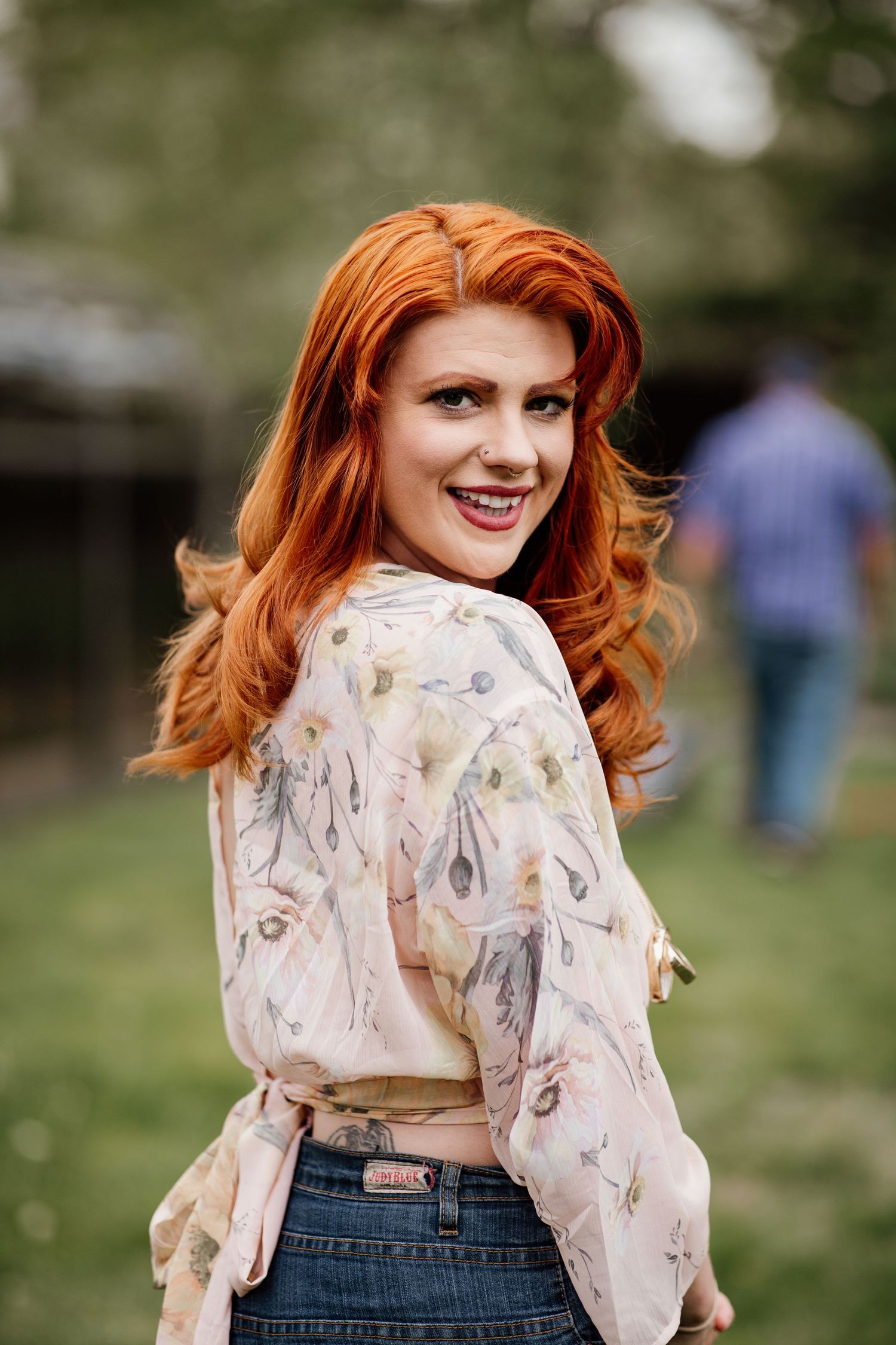 A smiling redhead woman in a floral blouse glances back over her shoulder outdoors.