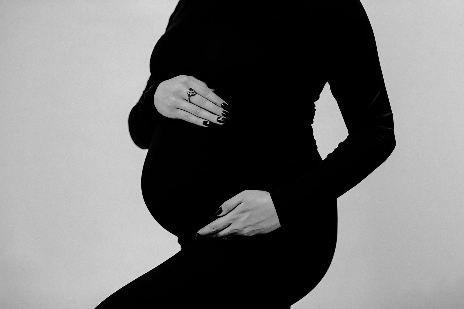 Black and white photo of a pregnant woman in a black outfit cradling her baby bump with both hands.