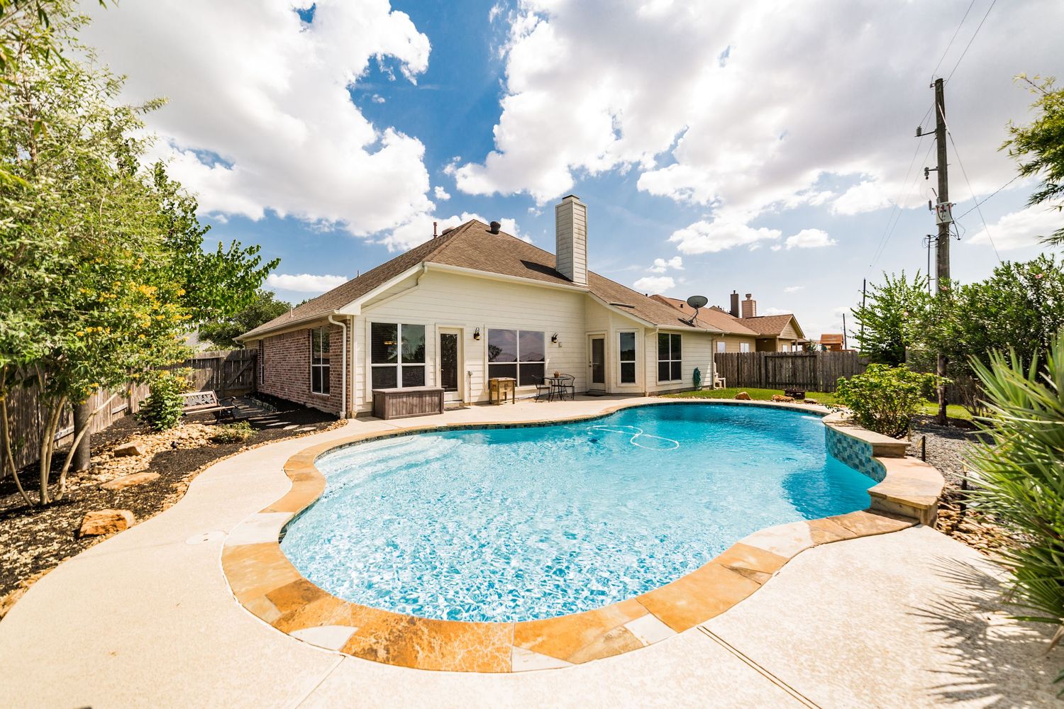 Backyard oasis featuring a freeform swimming pool with tan stone coping, lush landscaping, and a brick home with patio.