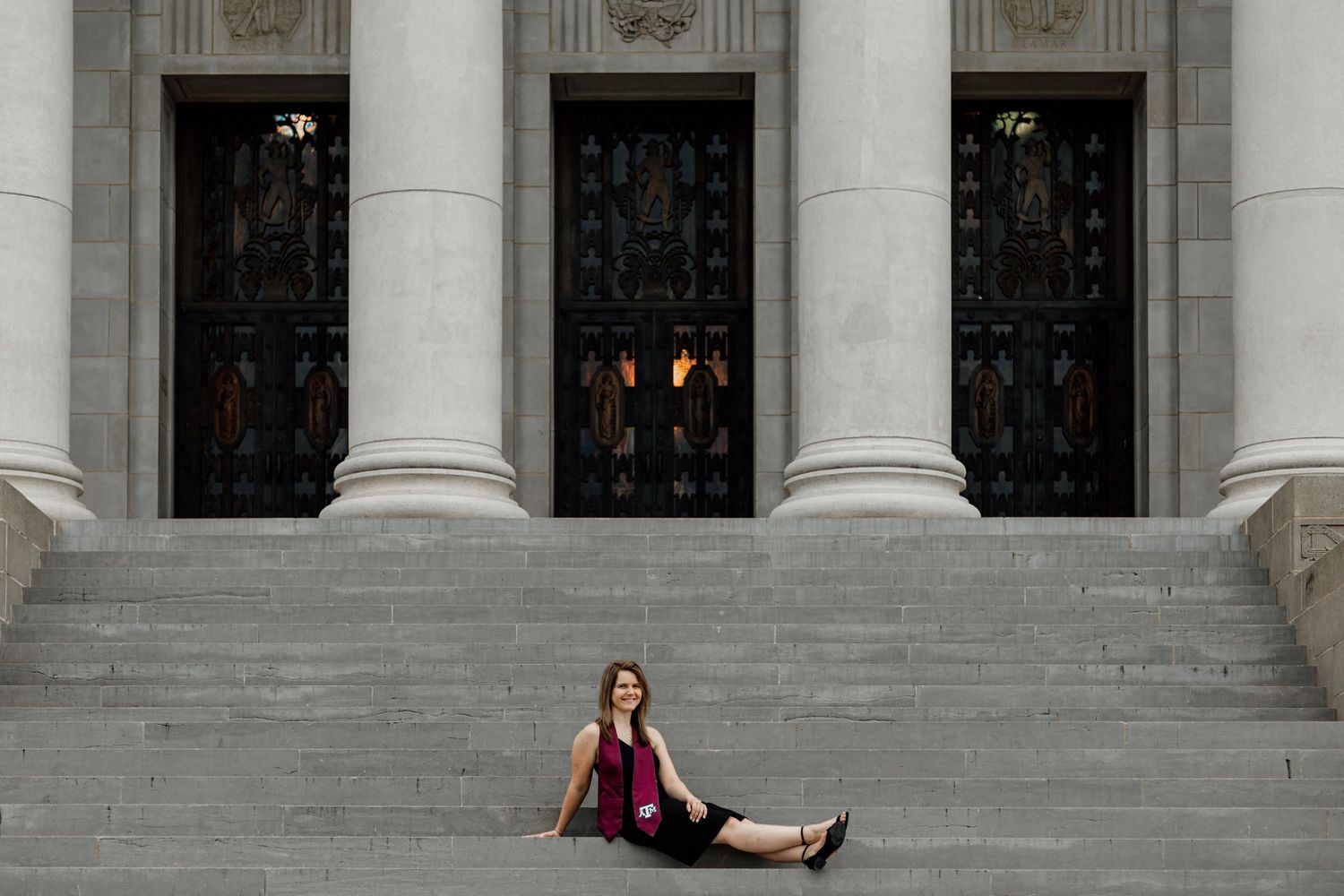 Woman in magenta top sits on steps of grand neoclassical building with ornate bronze doors and columns.